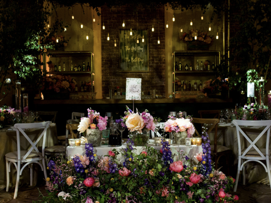 Decorated banquet table with pink, purple, and white flowers, set for a celebration in a dimly lit venue with hanging Edison bulbs, a bar in the background, and a floral arrangement at the center.