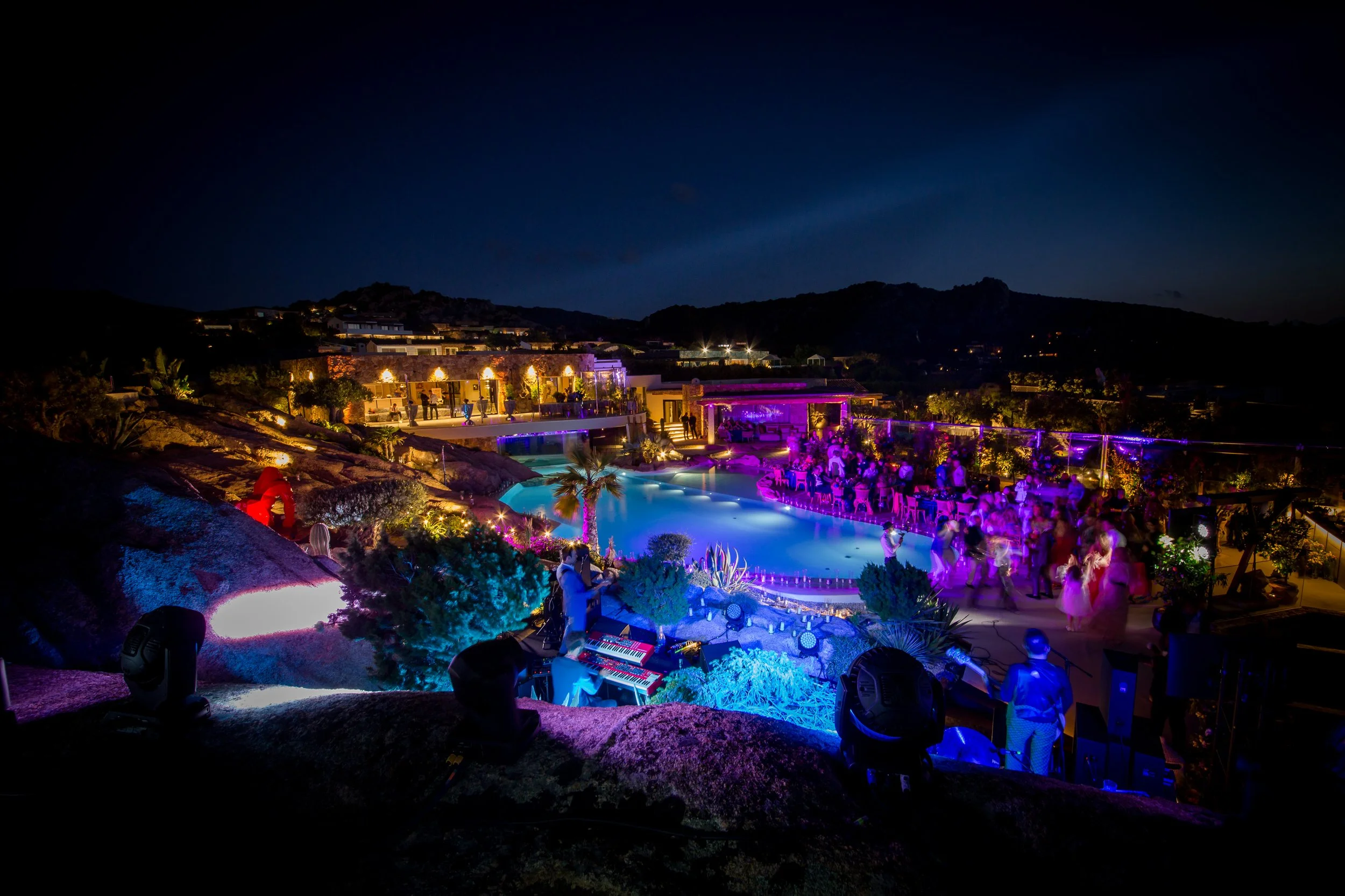 A Sardinia private birthday party at nighttime. This Villa outdoor party has an illuminated swimming pool, colorful lights, and guests dancing and socializing.