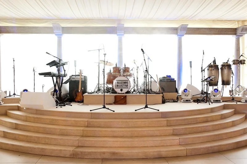 Empty stage with musical instruments, microphones, and lighting equipment, set up for a performance, with bright natural light coming from large windows in the background.