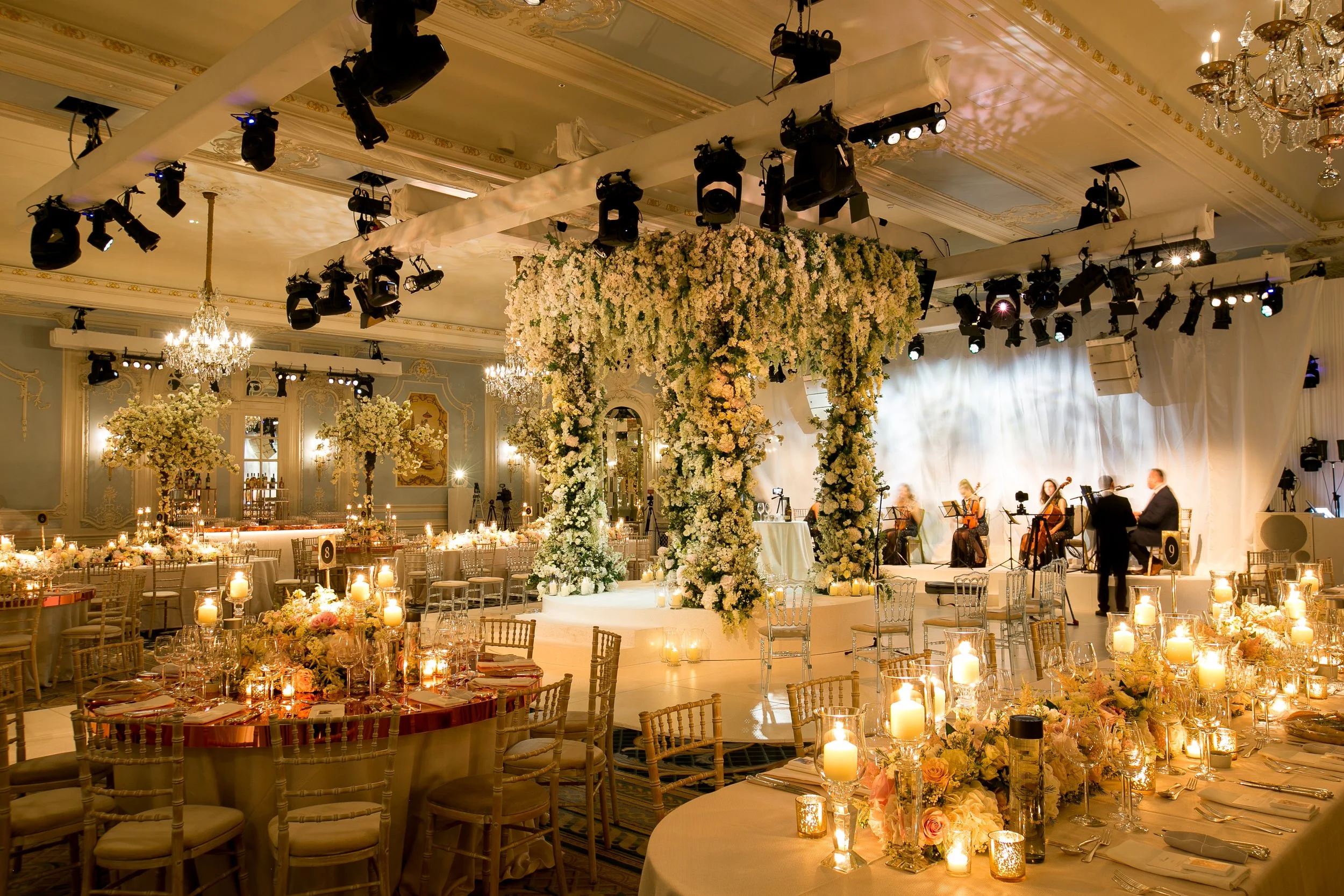 An elegant, decorated event hall with floral arrangements and candles on tables, with a stage hosting musicians and various lighting equipment hanging from the ceiling.