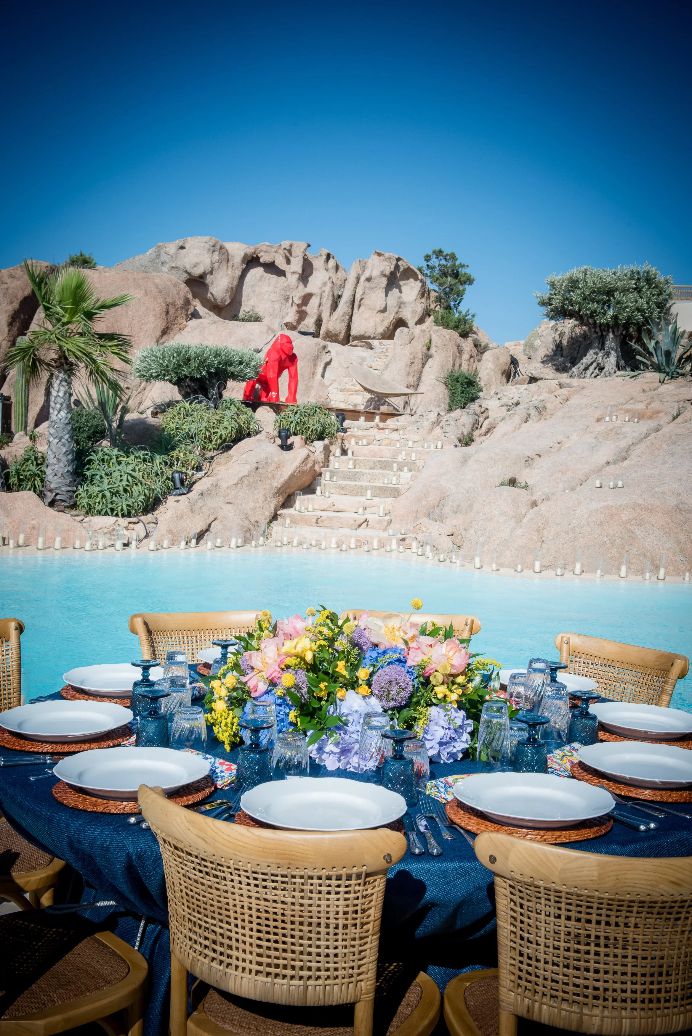 Outdoor dining area with a large round table set with white plates, glasses, and framed floral centerpiece, surrounded by woven chairs, overlooking a pool with decorative candles and a rocky desert landscape with trees and plants in the background.