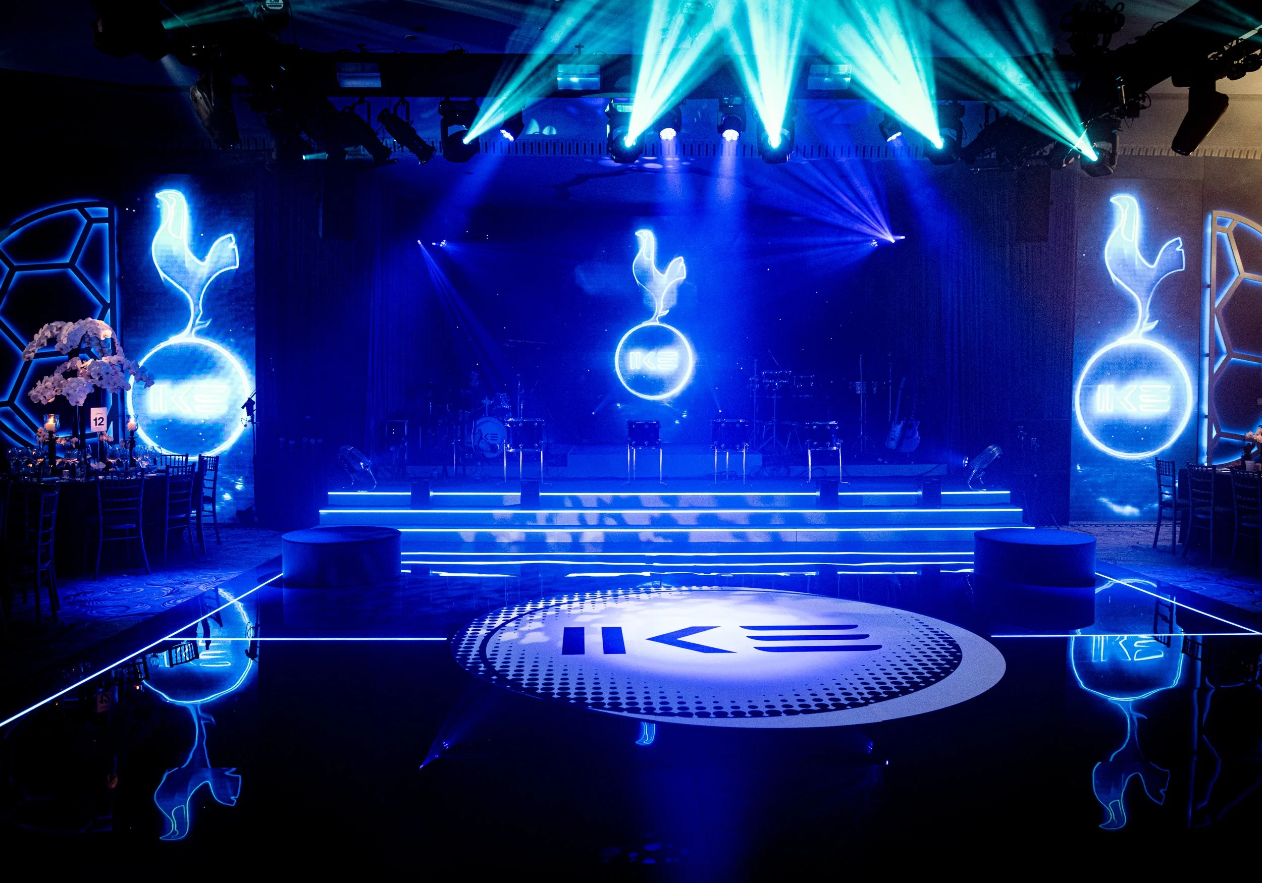 Decorated event stage with blue lighting, neon dolphin and flame logos, and illuminated circular floor logo, with seating and floral arrangements in the foreground.