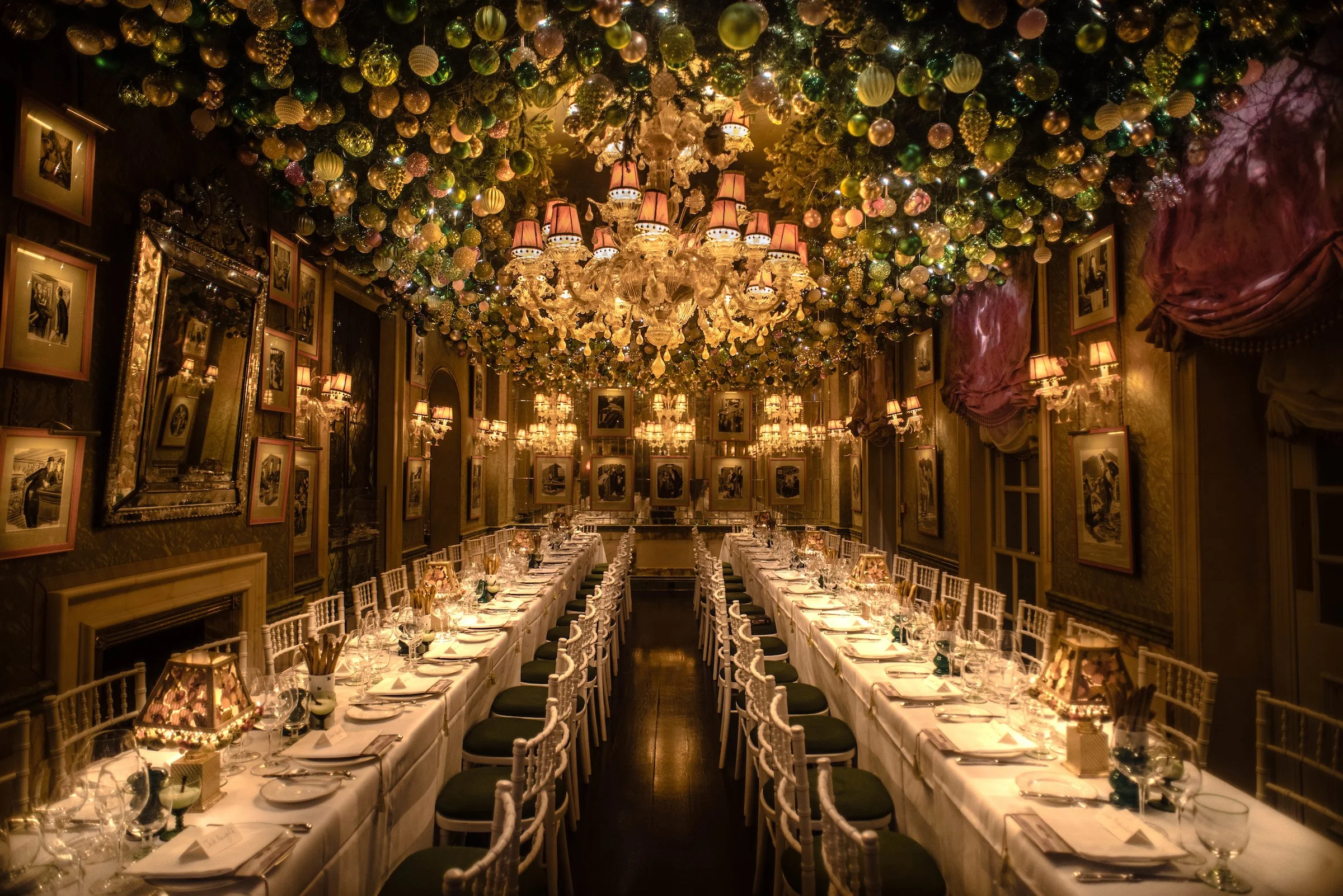 Elegant banquet room decorated for Christmas with a ceiling covered in gold and green ornaments, luxurious chandeliers, and framed black-and-white photos on the walls. Long tables are set with white tablecloths, candles, and place settings.