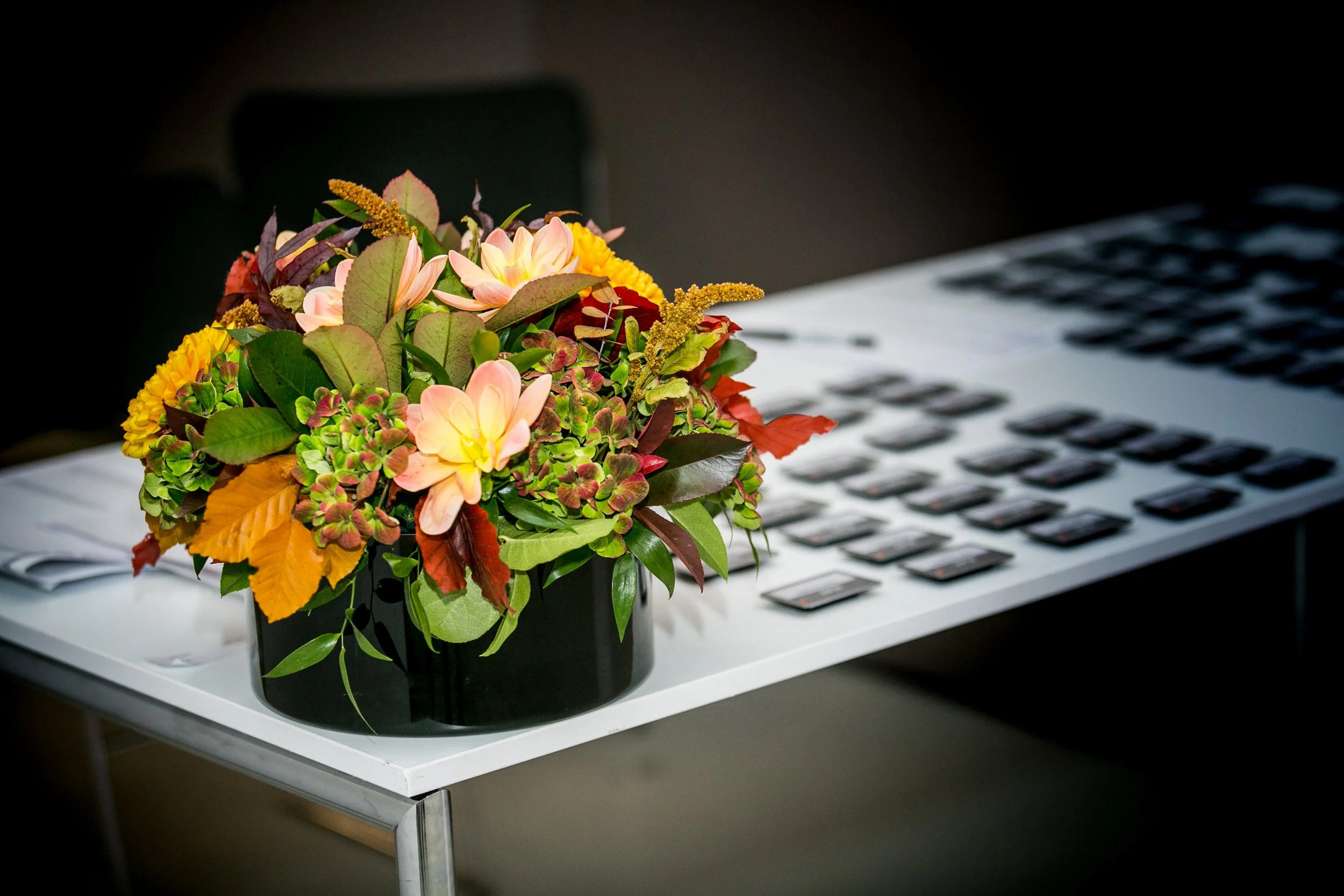 A bouquet of colorful flowers in a black vase on a white table next to a keyboard.