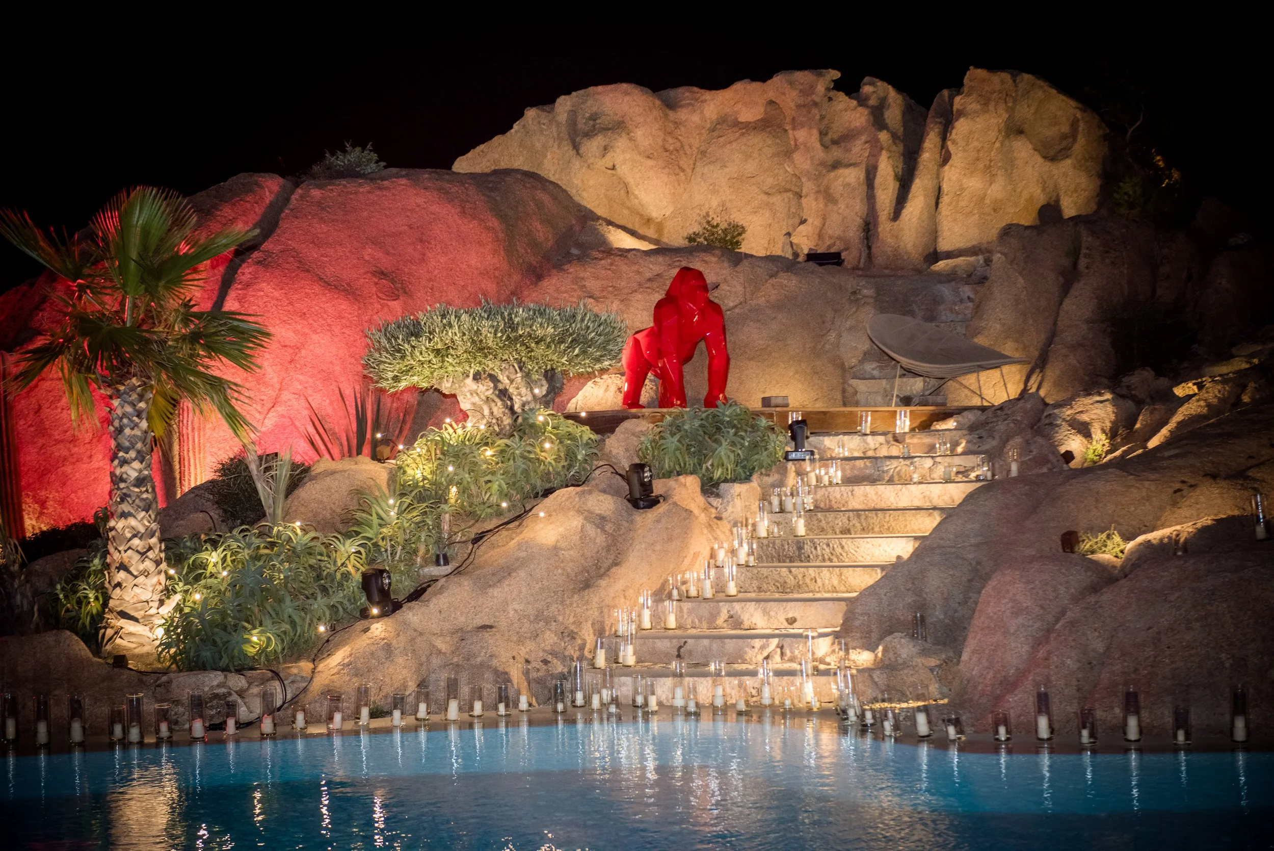 Nighttime scene of illuminated pool with candles, rocky landscape, and desert plants, including a prominent red sculpture resembling a dog.