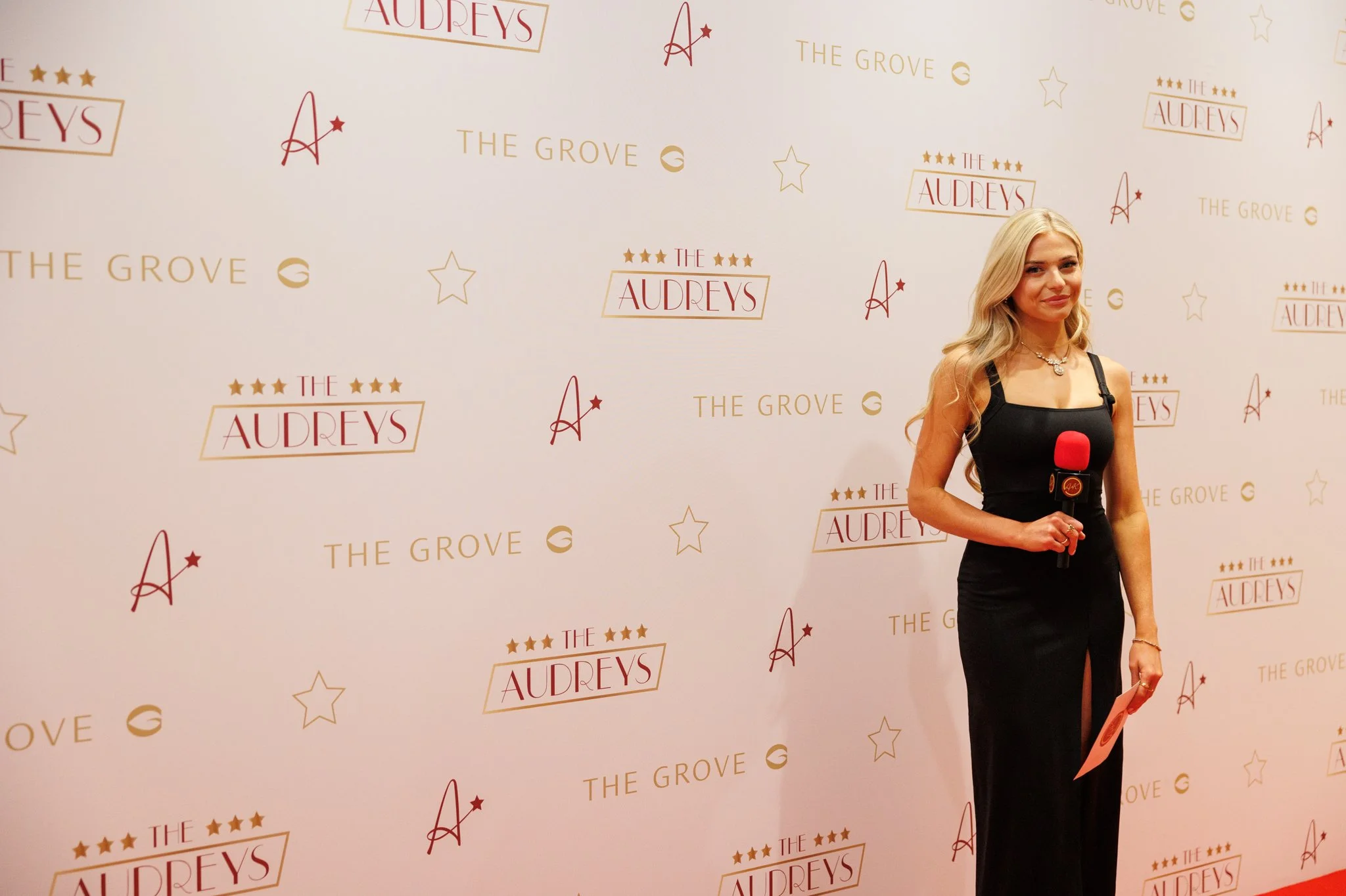 A woman with long blonde hair wearing a black dress with a thigh-high slit and holding a microphone stands in front of a step-and-repeat backdrop with logos for "The Audreys" and "The Grove" decorated with stars.
