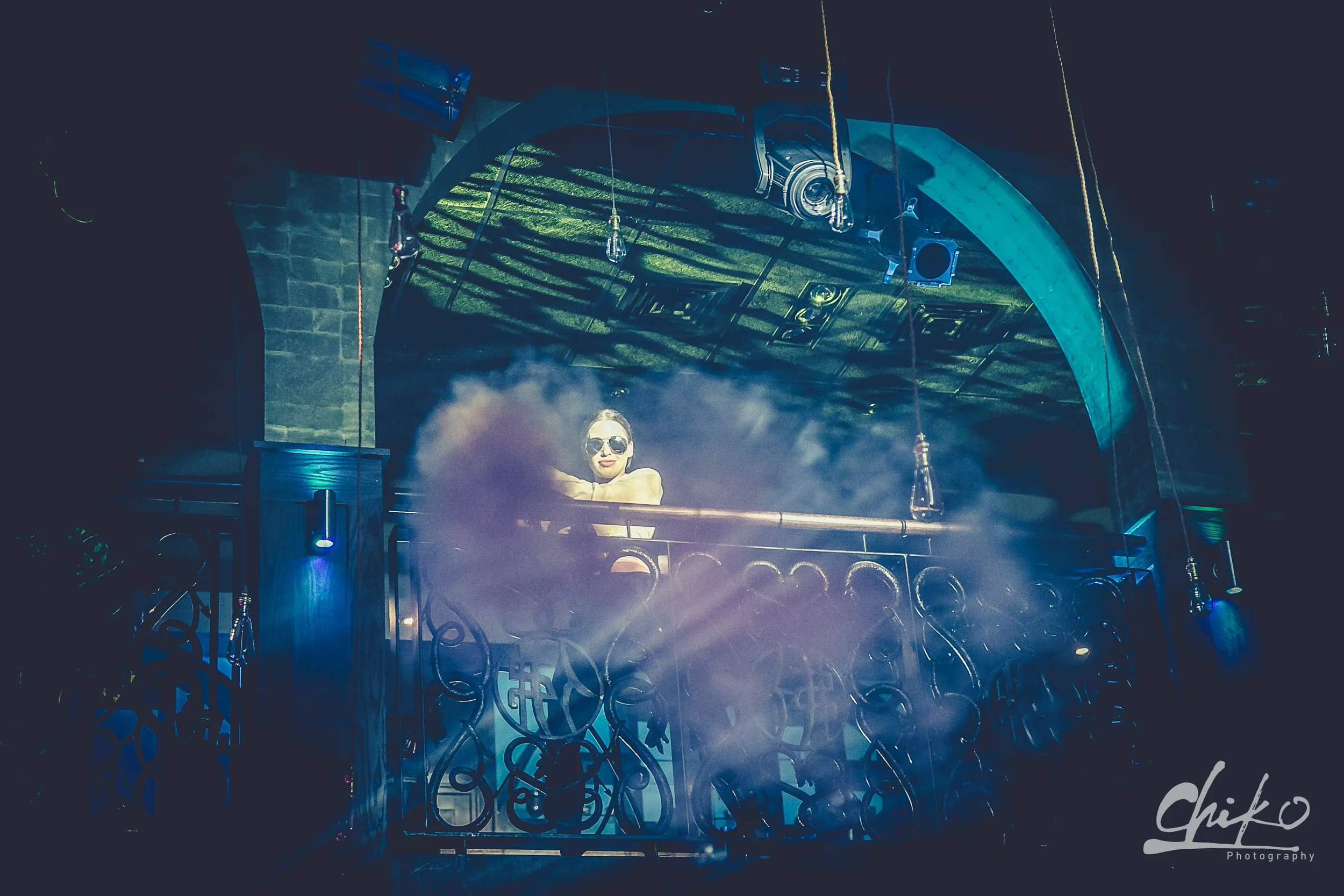 A person with sunglasses standing behind a DJ booth on a stage, with stage lights and equipment hanging above, in a dark club or concert setting.