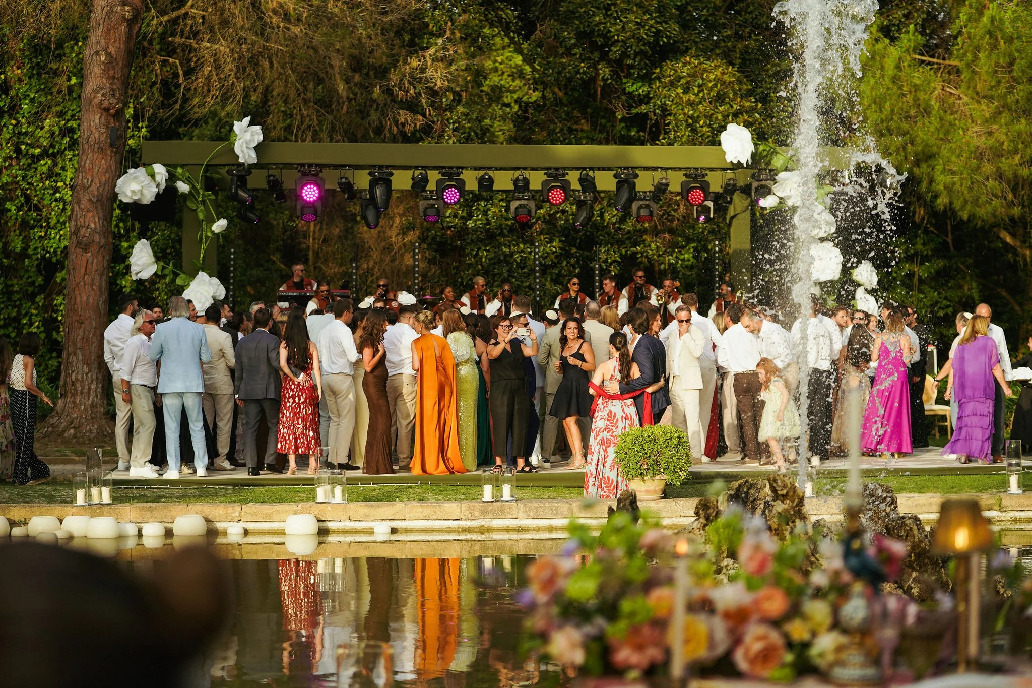 A large gathering of people in formal attire at an outdoor event near a fountain, with a stage and a band performing, surrounded by trees and greenery at Anantara Villa Padierna Resort in Marbella