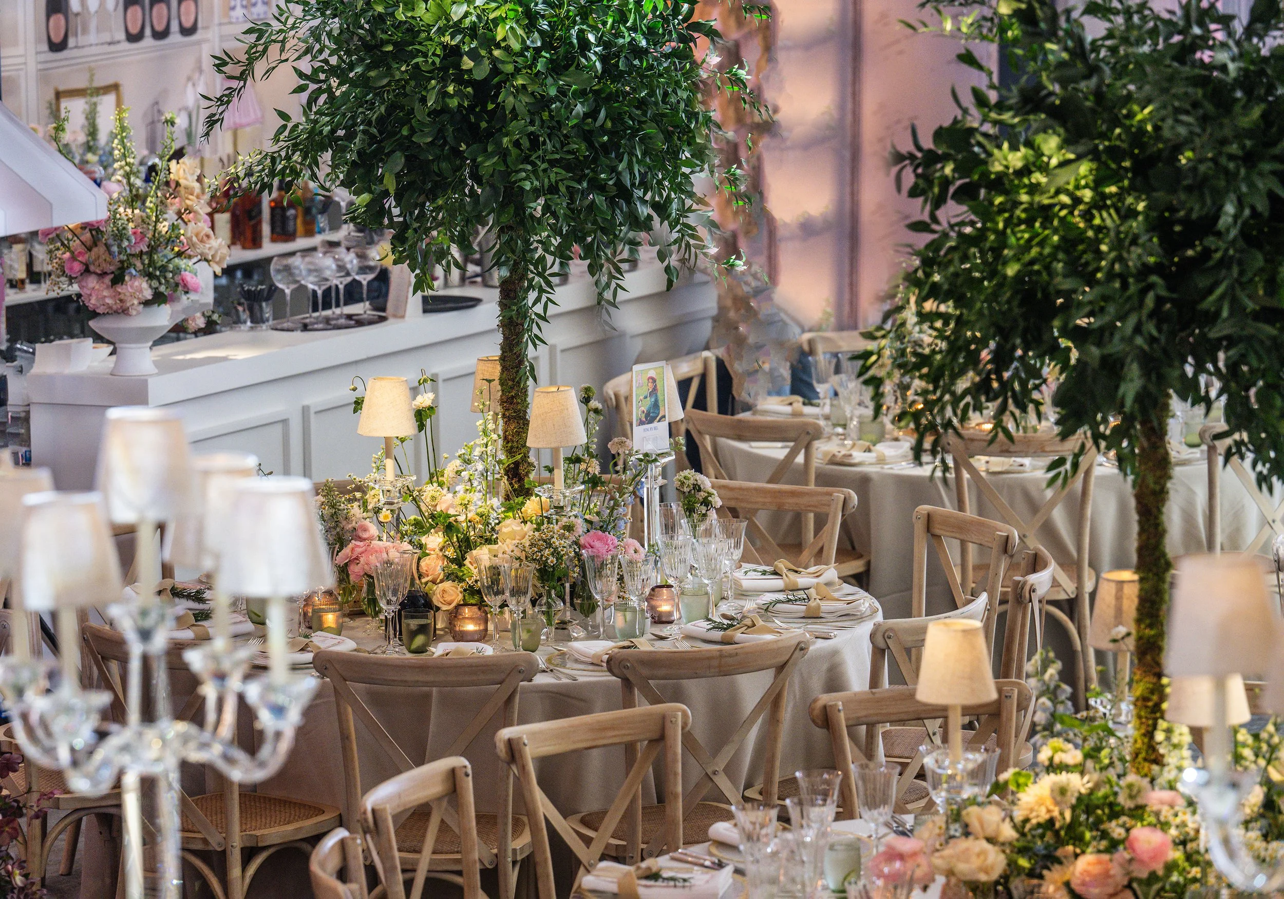 A decorated dining area for a Bat Mitzvah, featuring long tables with floral centerpieces, candles, and table settings, surrounded by chairs, with large green trees and soft lighting creating an elegant atmosphere.