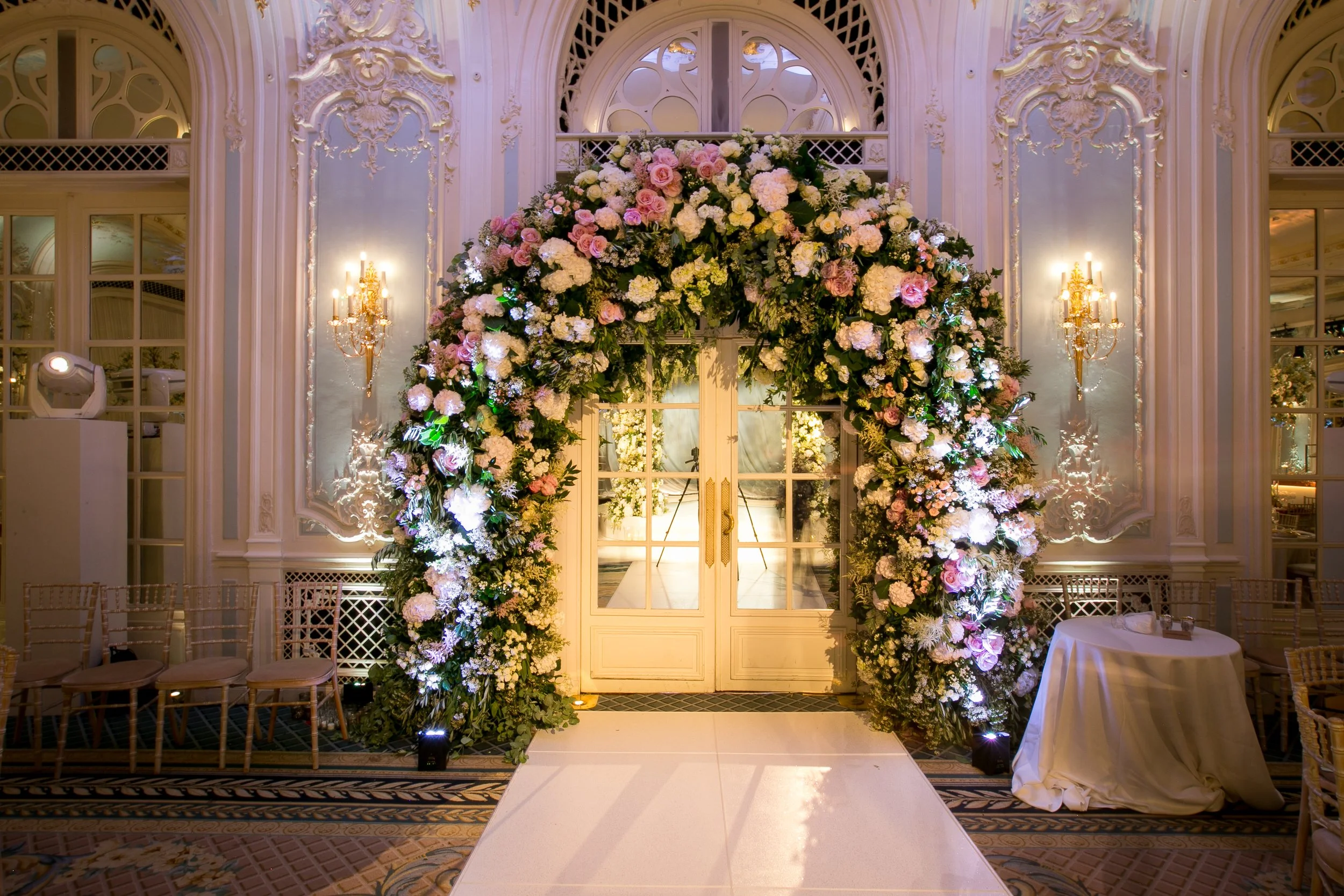 An elegant wedding altar decorated with a large floral arch composed of white and pink flowers inside a lavishly decorated room with ornate white walls, gold fixtures, and glass doors.