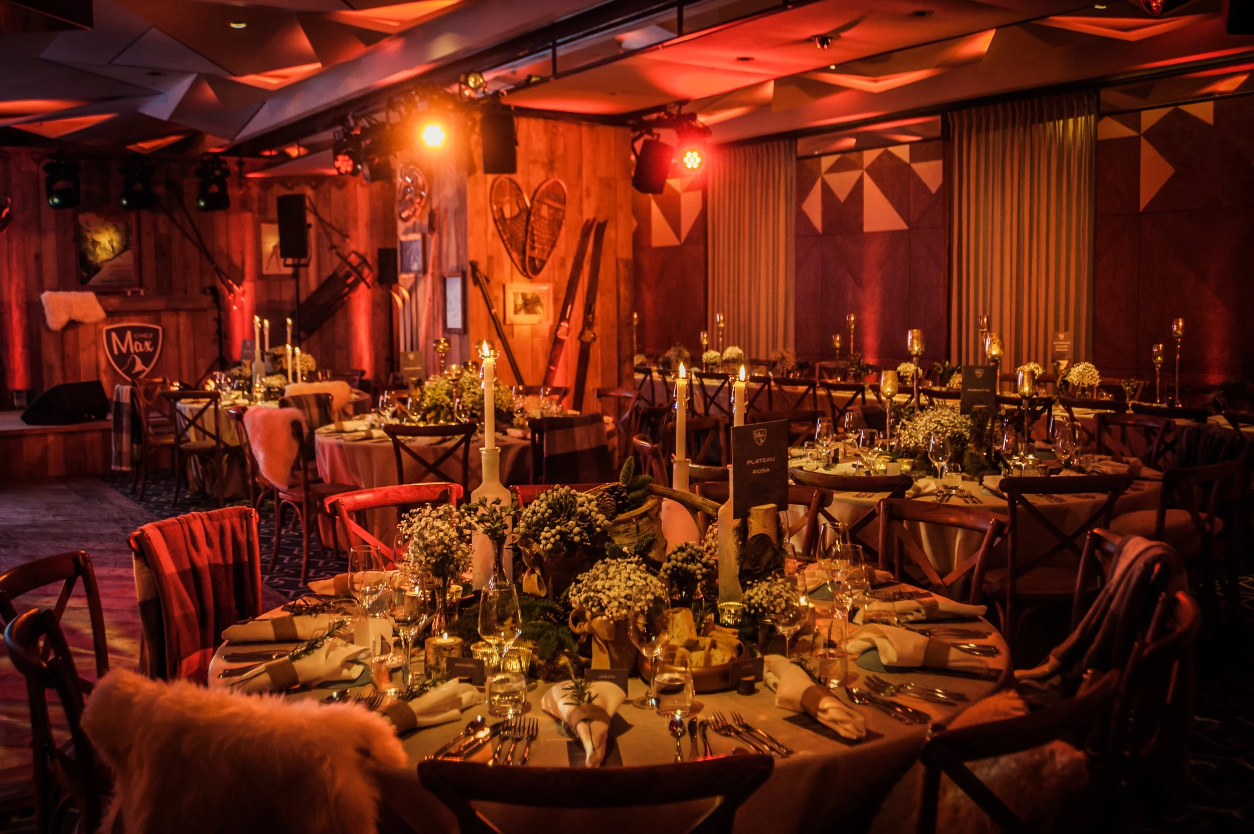 A decorated banquet hall with round tables set for a formal dinner, featuring floral centerpieces, candles, wine glasses, and tableware. The room has wooden walls, wall art, and warm lighting, with a stage or performance area at the front.