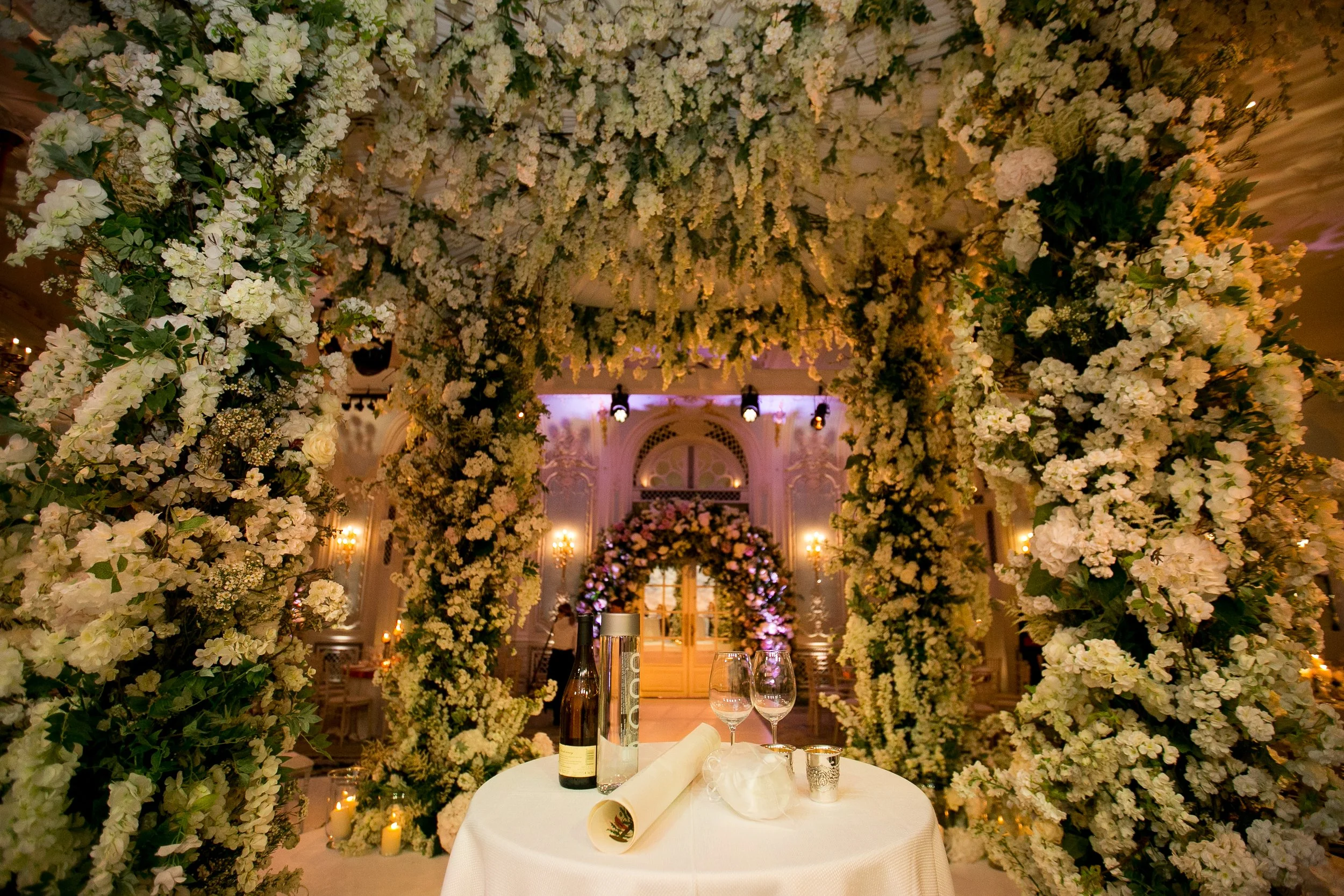 A decorated wedding reception space with a floral archway made of white flowers and greenery, a table set with wine bottles, wine glasses, candles, and tableware, and an ornately decorated interior with chandeliers and draped fabric in the background