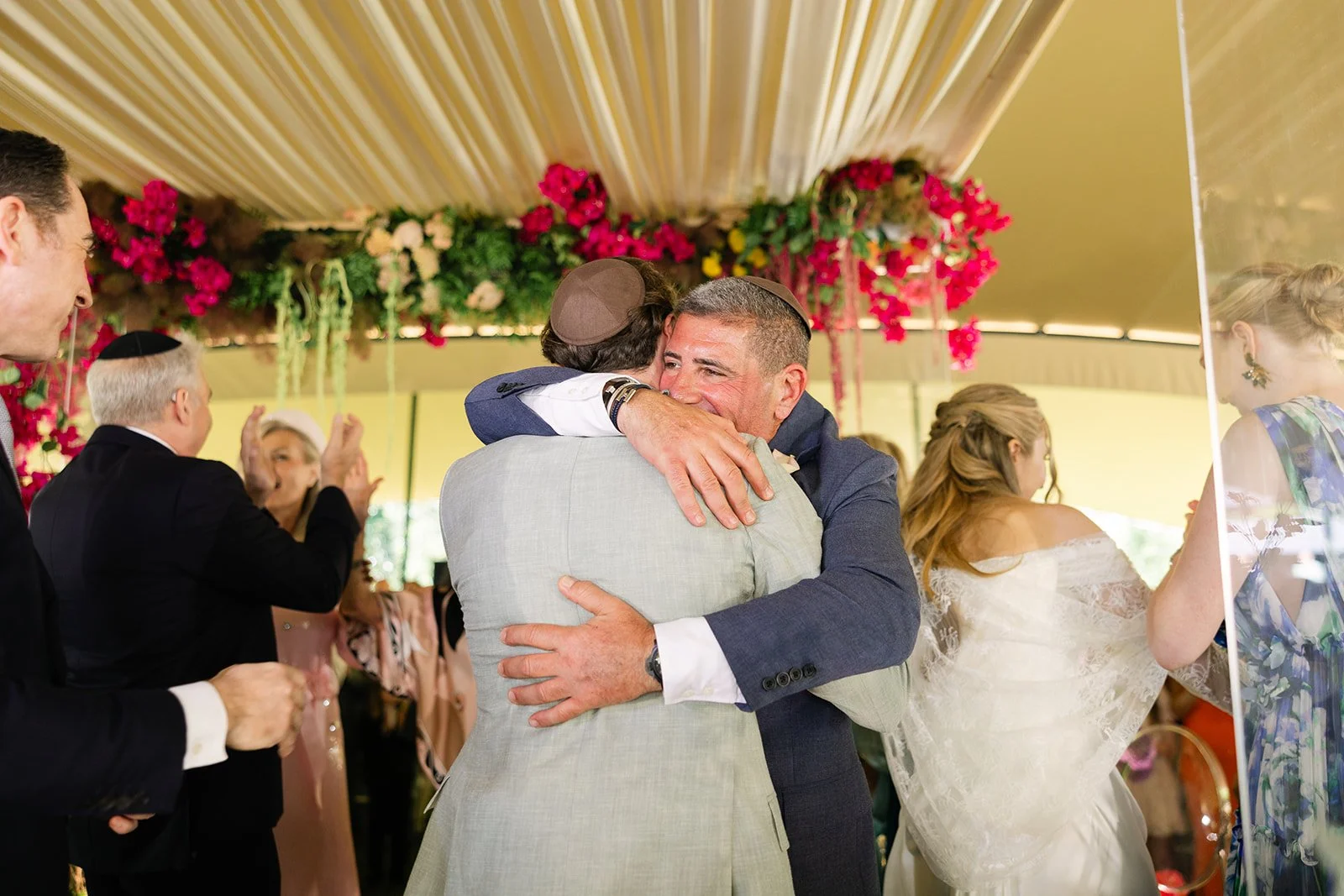 Two men embrace and hug at a celebration, with one wearing a kippah. People are clapping and smiling around them, with a decorated floral backdrop.