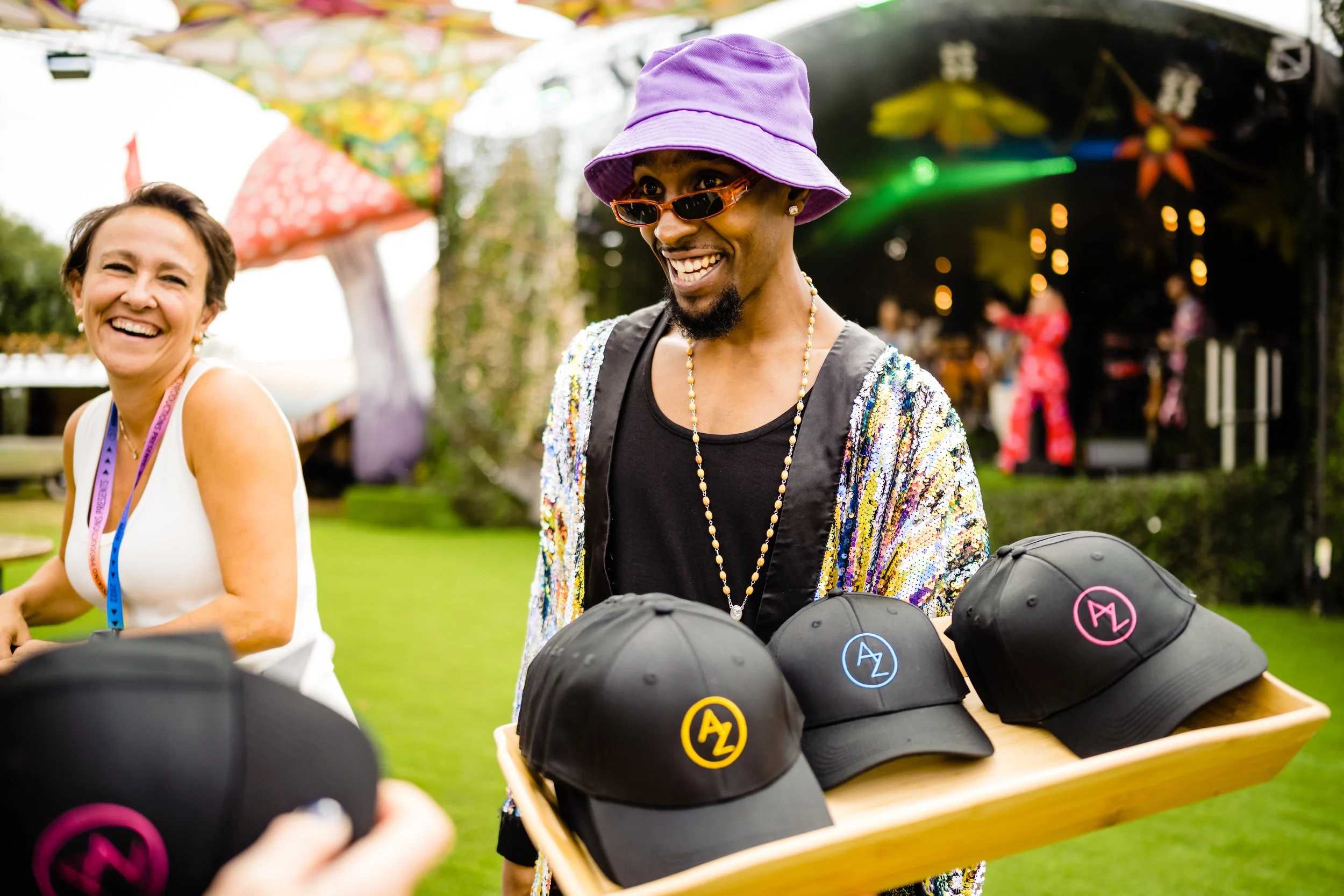 A smiling woman wearing a white top and purple lanyard on the left, and an African American woman wearing a purple hat, sunglasses, colorful sequin jacket, and necklace on the right, standing outdoors at a daytime event with hats on a table in the fo