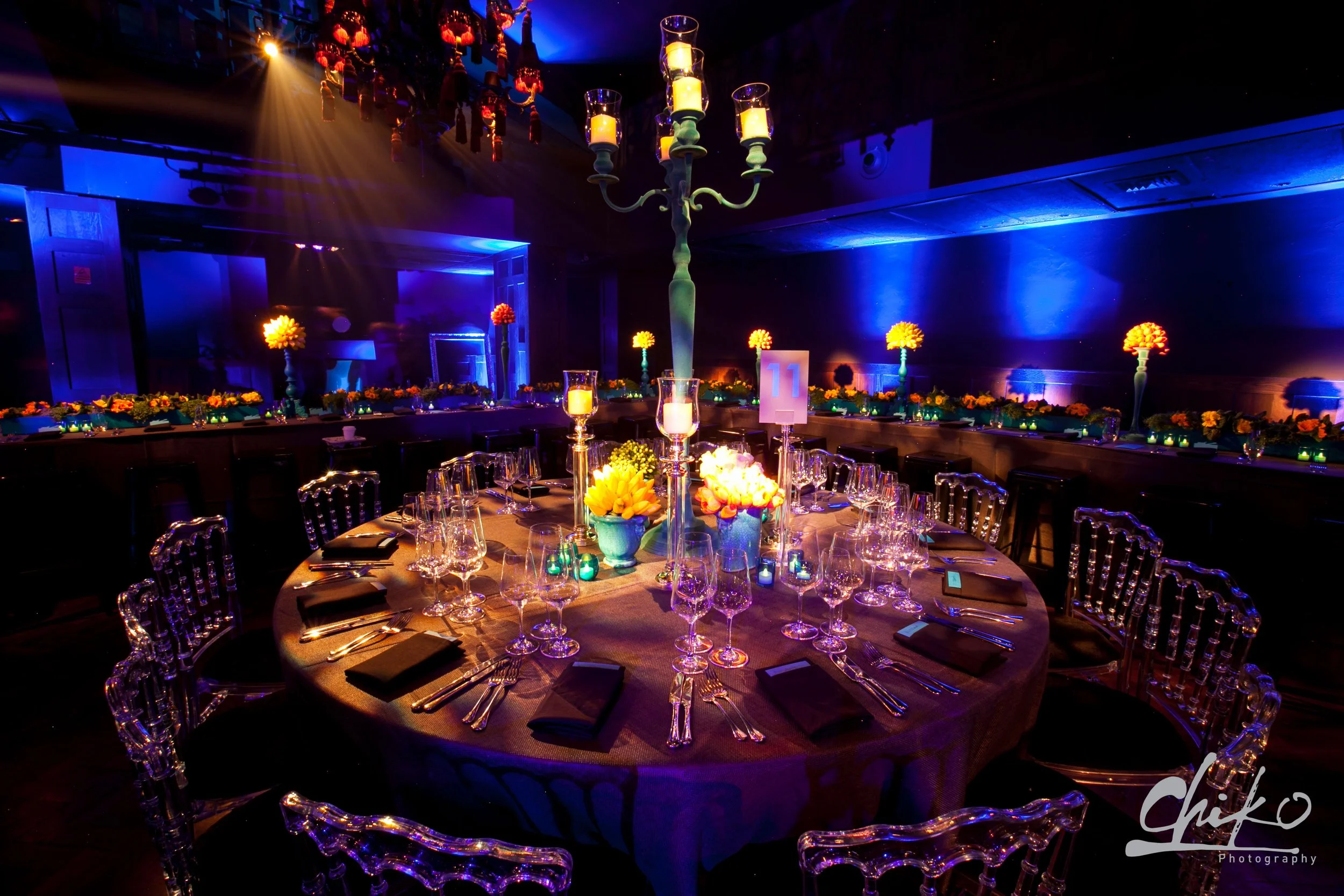 Elegant banquet table set for a formal event, decorated with flowers, candles, and glassware, in a dimly lit room with blue and yellow lighting and a chandelier.