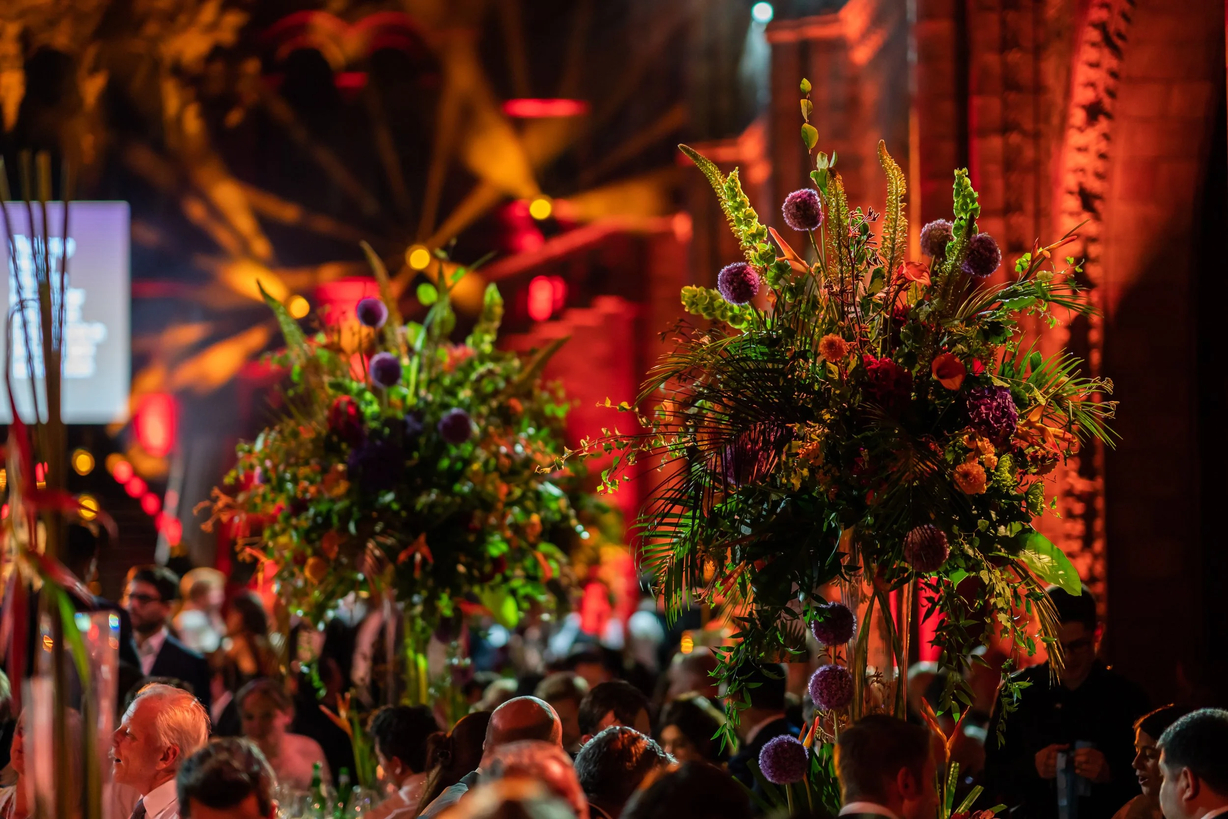 A crowd of people attending a corporate event, with large floral arrangements featuring purple globe flowers and green foliage, illuminated by colorful stage lights at The Natural History Museum