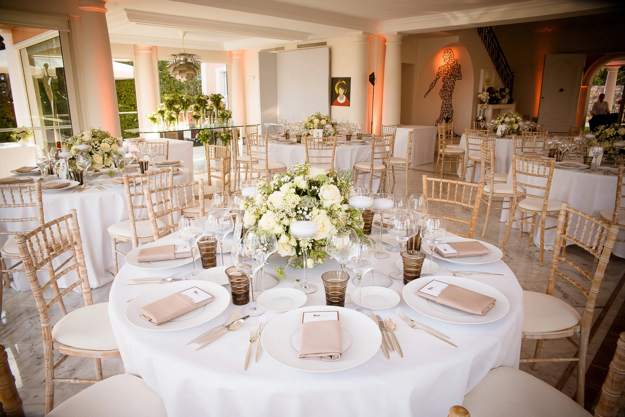 Elegantly decorated banquet hall with round tables covered in white tablecloths, each set with plates, silverware, wine glasses, and beige napkins. Centerpieces feature white flowers and greenery. Chiavari chairs surround each table. Natural light st