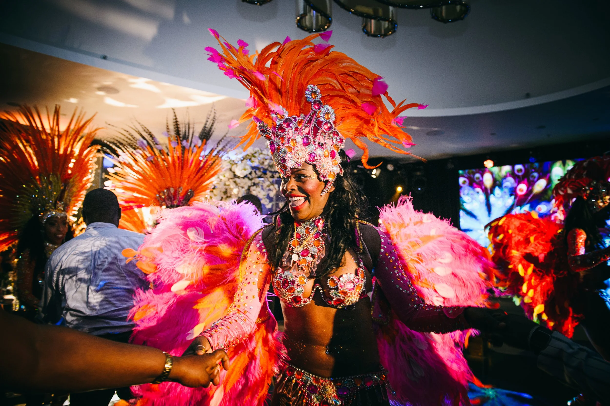 A woman dressed in a vibrant carnival costume with pink, orange, and purple feathers, jewelry, and a large feathered headdress, smiling during a parade or celebration.