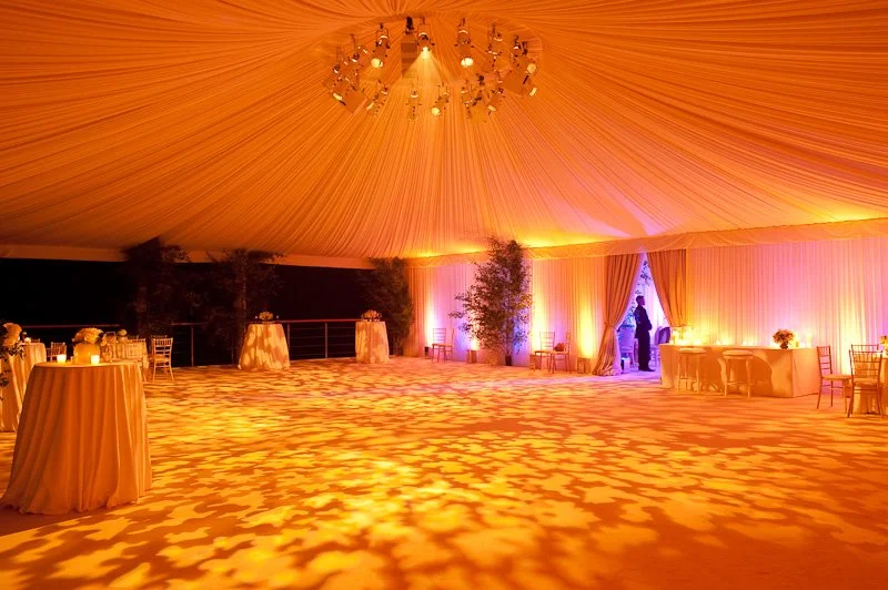 Elegant event space under a large orange draped ceiling with decorative lighting, tables with white tablecloths, and plants, illuminated with warm lighting.