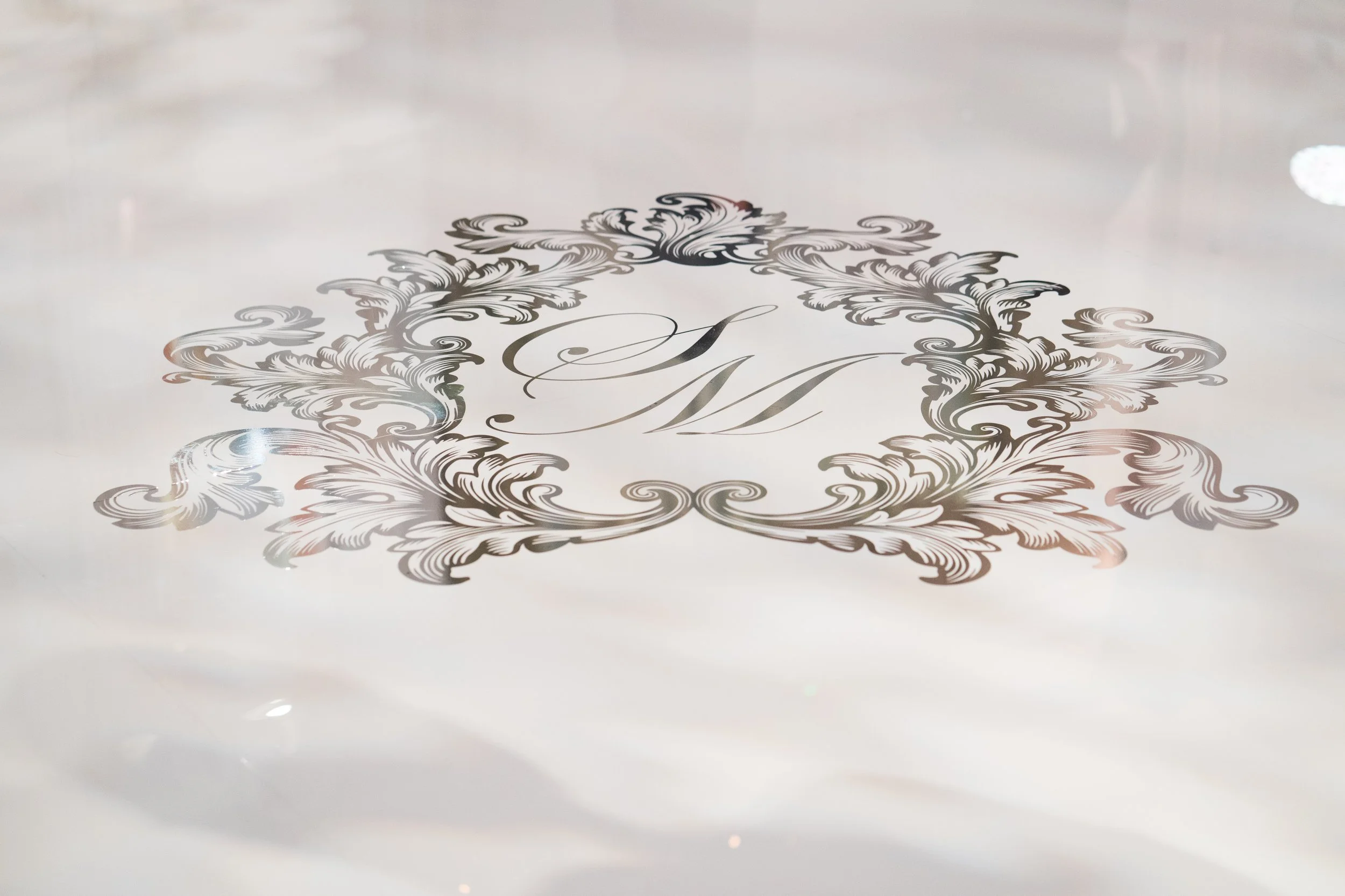 Close-up of a decorative emblem or monogram with ornate floral patterns and the letter M in the center, engraved on a smooth, reflective surface.