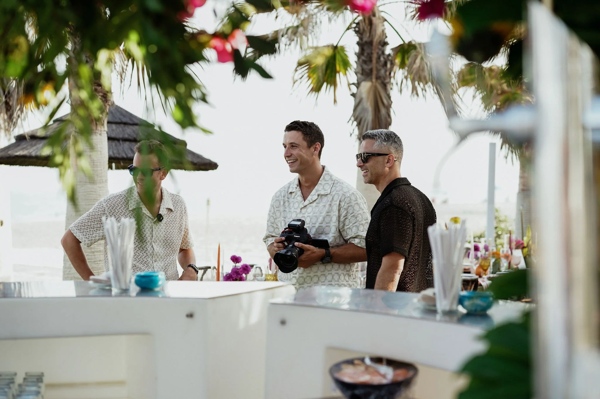 Just Seventy Stas Anastasiou and Laurie Weitzkorn and Reece Iveson standing together outdoors, smiling, and holding a camera, at a beachside event with tropical trees and decorated tables.