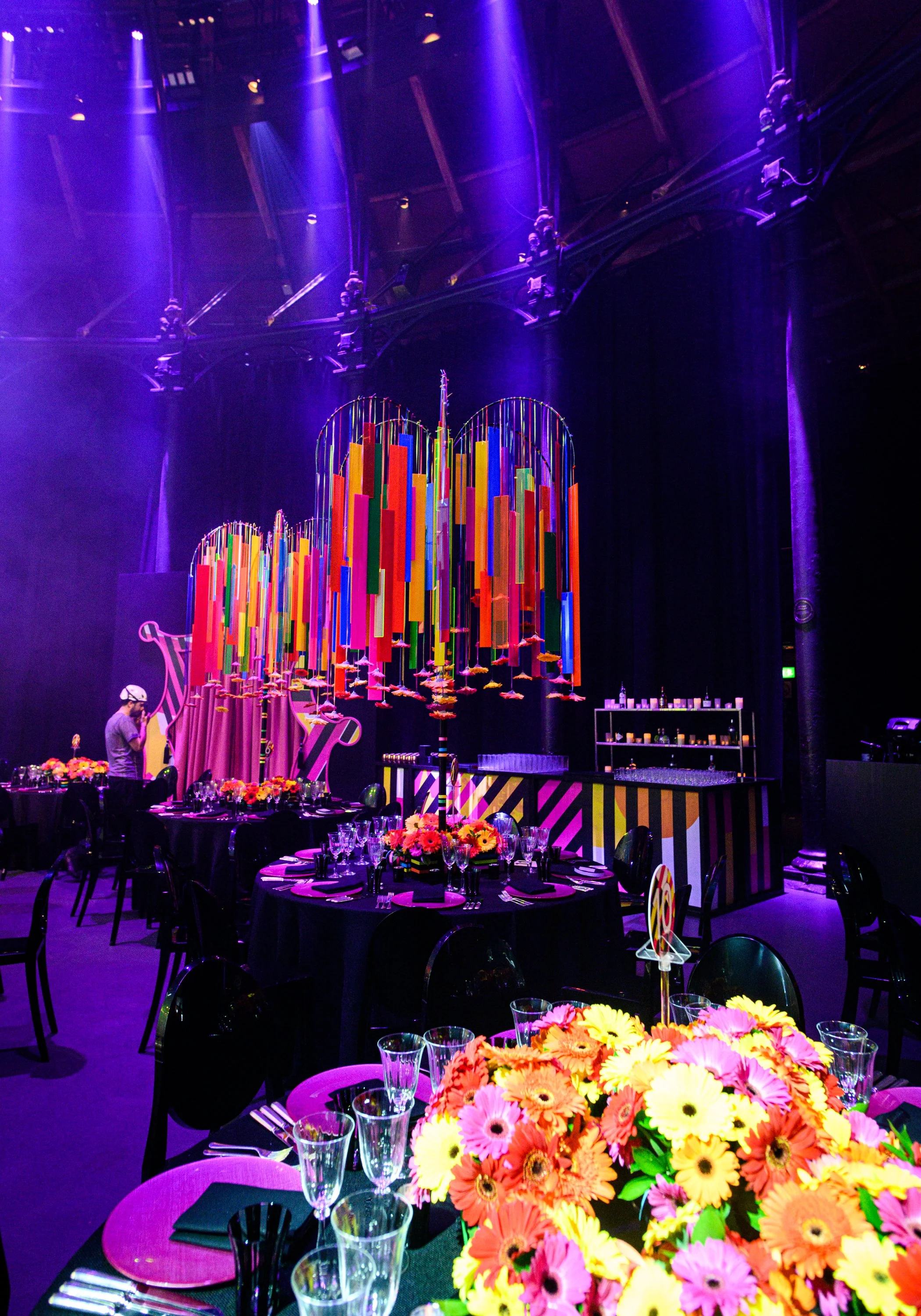 Colorful floral centerpiece with orange, yellow, and pink flowers on a table set for a formal event with black chairs, glassware, and dark tablecloths, under purple lighting in a venue with circular hanging light fixtures and stage area.  Bat Mizvah 
