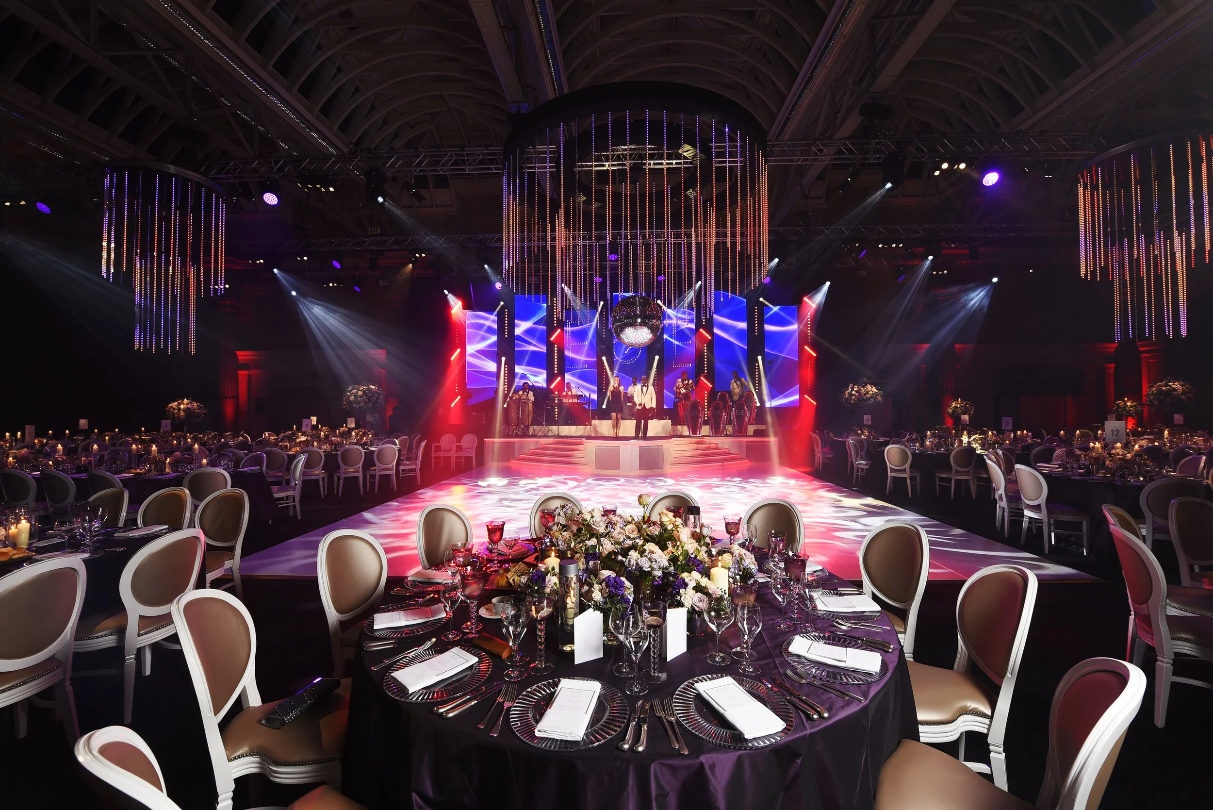 An elegant banquet hall decorated with round tables draped in purple tablecloths, set with glassware, cutlery, and floral centerpieces. The stage at the front features a band performing and a large disco ball hanging from the ceiling, with colorful lighting and vertical LED light strips hanging from the ceiling.