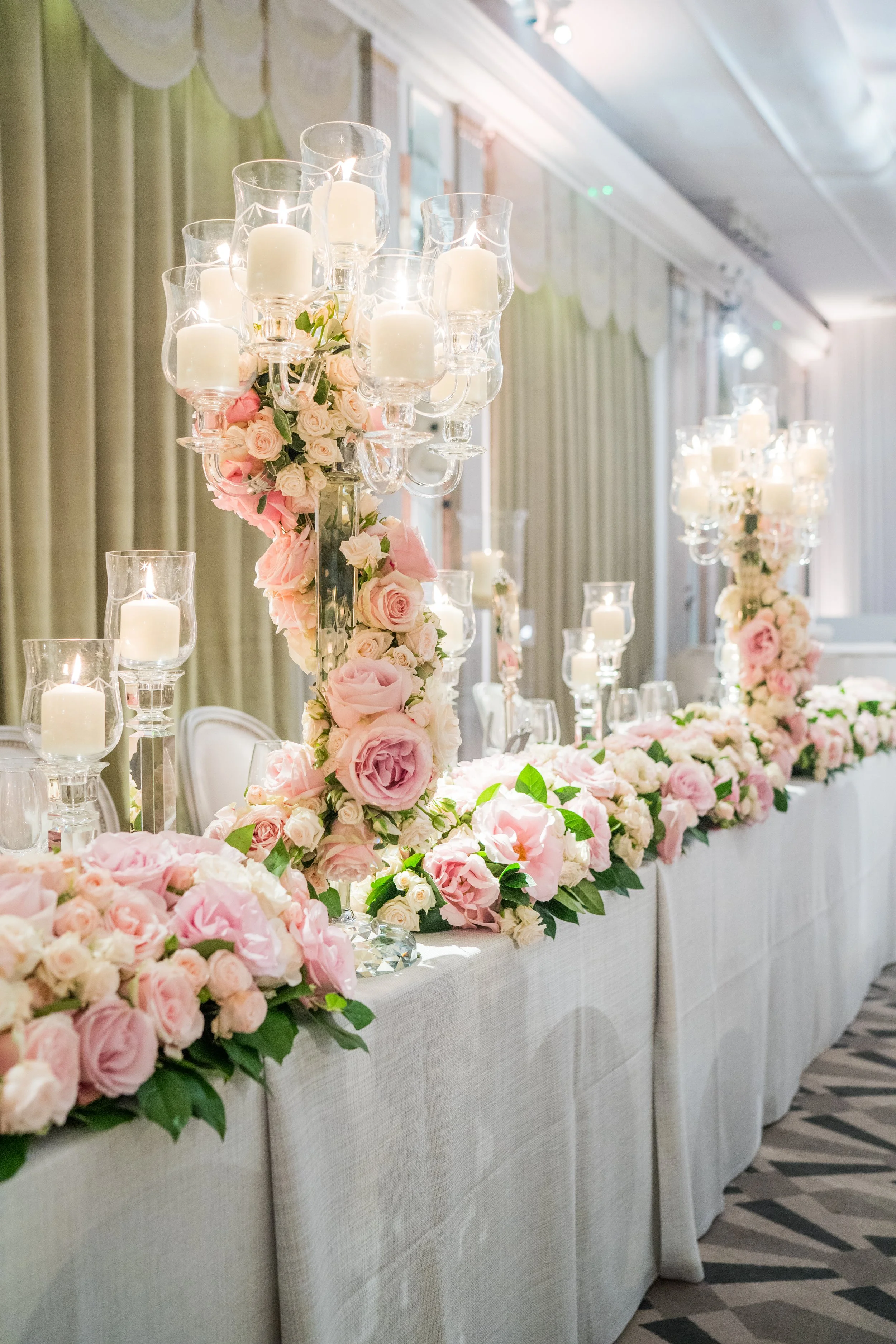 Elegant banquet table decorated with tall crystal candle holders filled with white candles, surrounded by pink and white roses and greenery, in a room with draped curtains and patterned carpet.