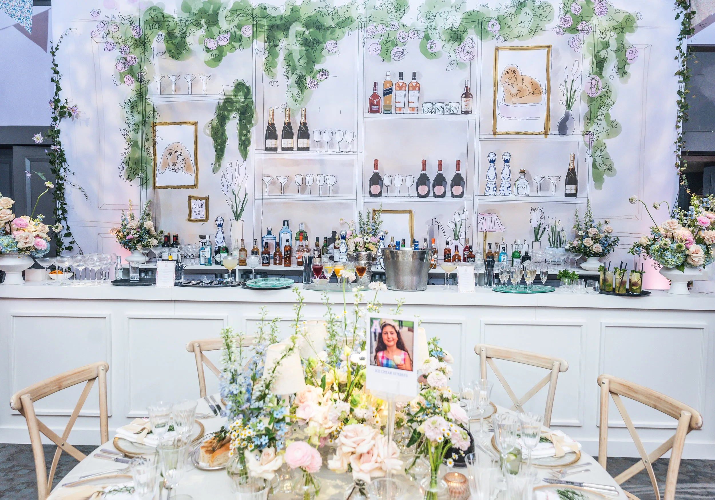 A decorated party backdrop with floral arrangements, a bar with bottles of alcohol, glassware, and framed dog pictures. In front, there are tables with floral centerpieces and place settings, set for a Bat Mitzvah at Grosvenor House on Park Lane 