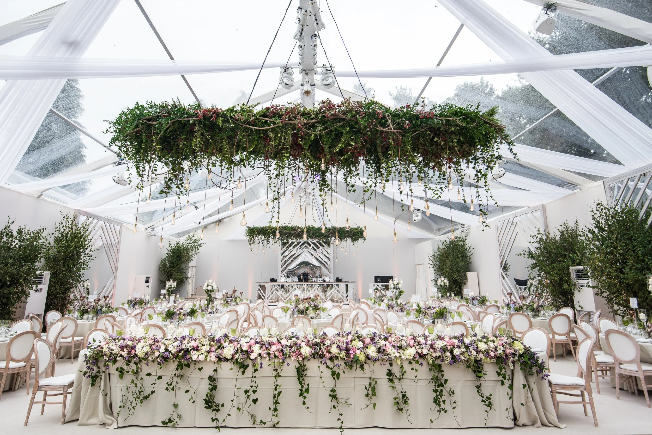 Elegant event venue decorated with floral arrangements, featuring a large floral centerpiece on a long table and hanging greenery ceiling installation with draped white fabric and string lights.