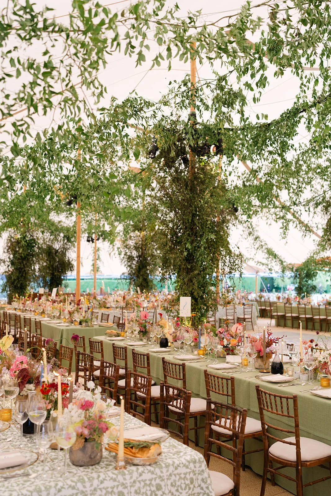 A large wedding reception setup inside a tent with long tables decorated with floral arrangements, candles, and tableware. A large artificial tree with green leaves stands in the center, and the background shows additional tables and chairs.