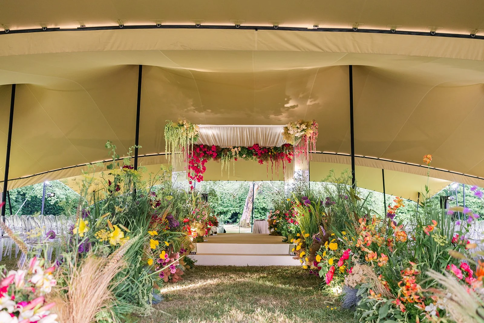 Decorated outdoor wedding chuppah with flowers, drapery, and seating under a large tent.