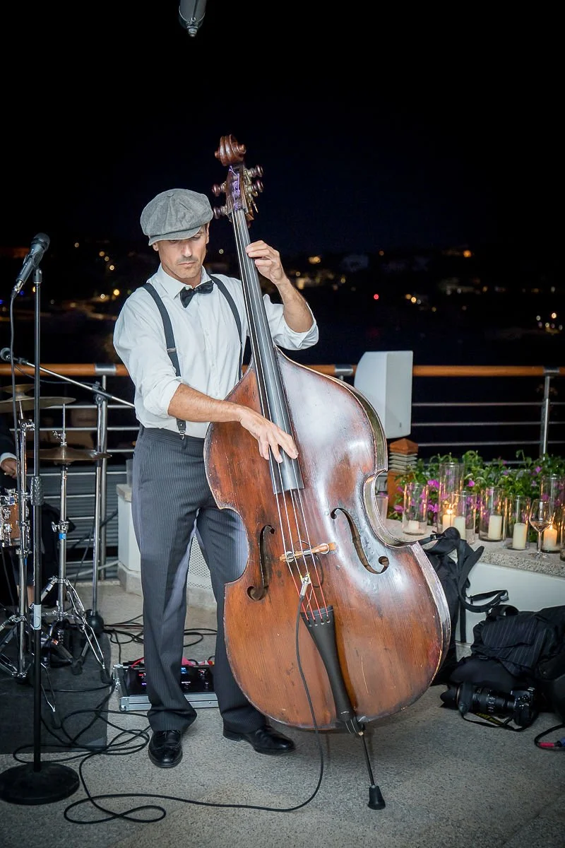 A man dressed in vintage attire, playing a double bass during a nighttime outdoor music event on a balcony with city lights in the background.