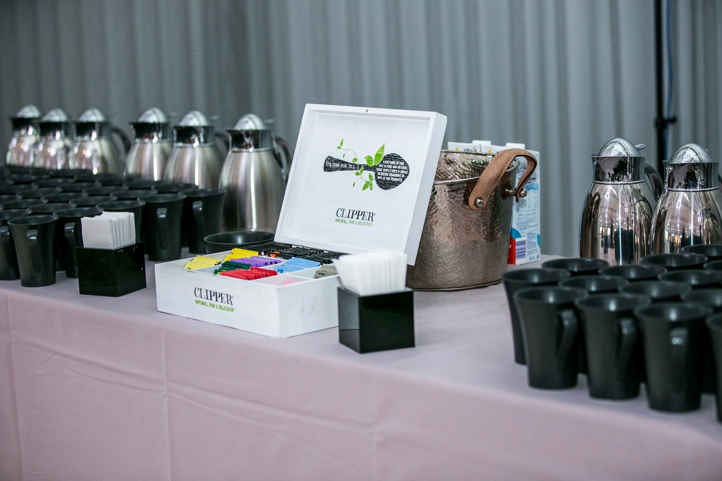 Table with coffee carafes, black cups, and a white box of various tea bags in front of a gray corrugated wall.