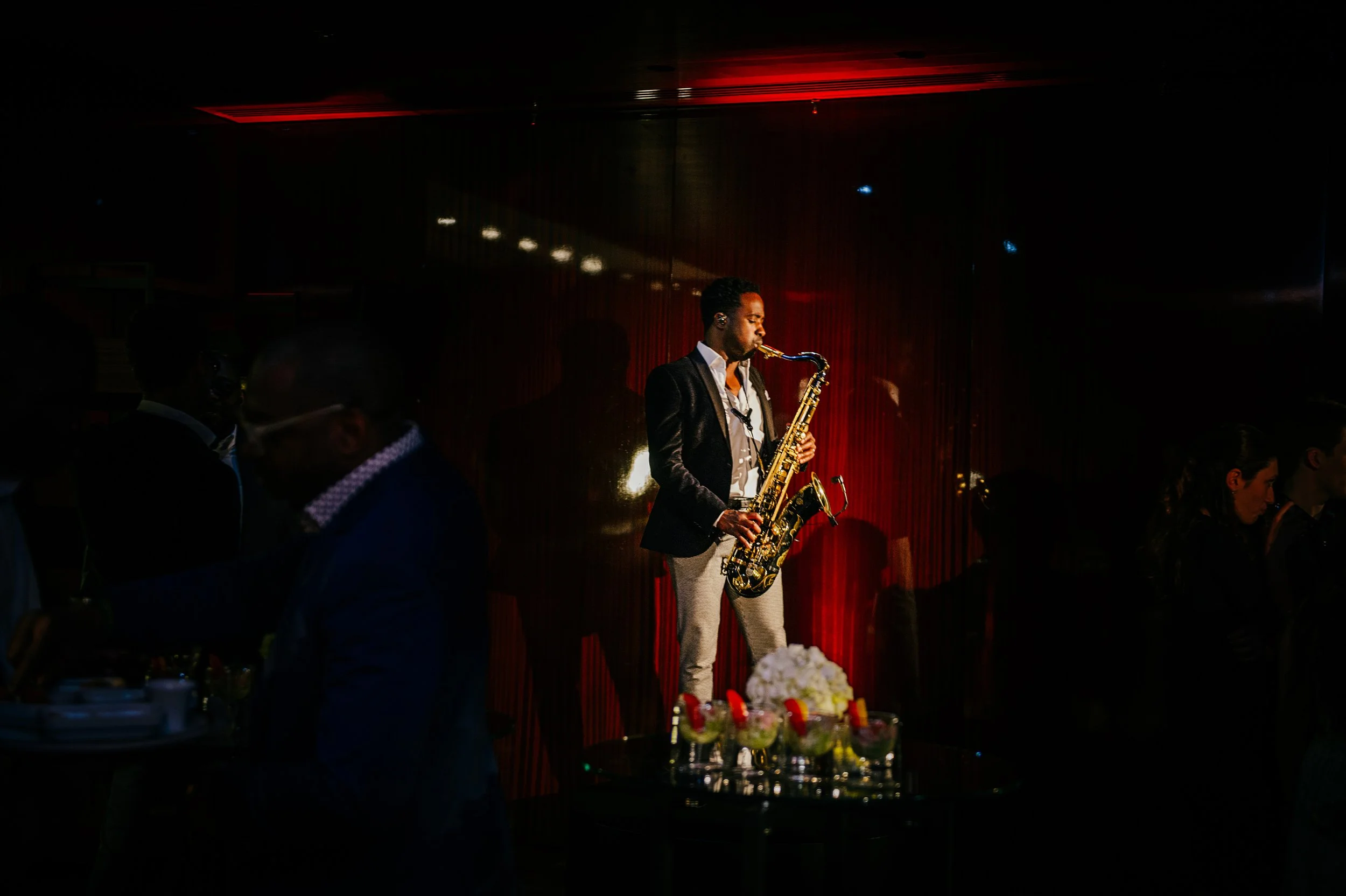A man playing a saxophone on a dimly lit stage with red curtains, surrounded by an audience at a formal event.