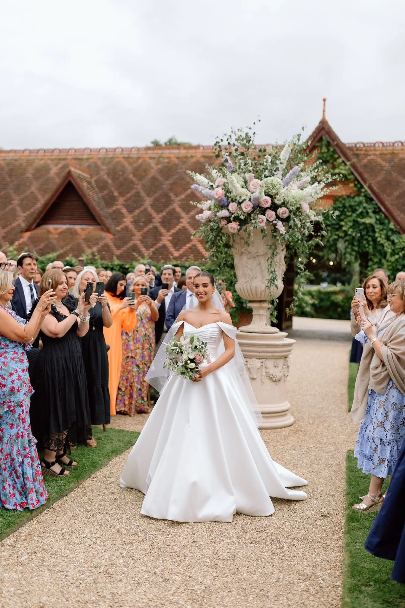 Bride in white wedding gown holding a bouquet, smiling, surrounded by guests taking photos at an outdoor wedding ceremony with a large floral arrangement and a brick building in the background.