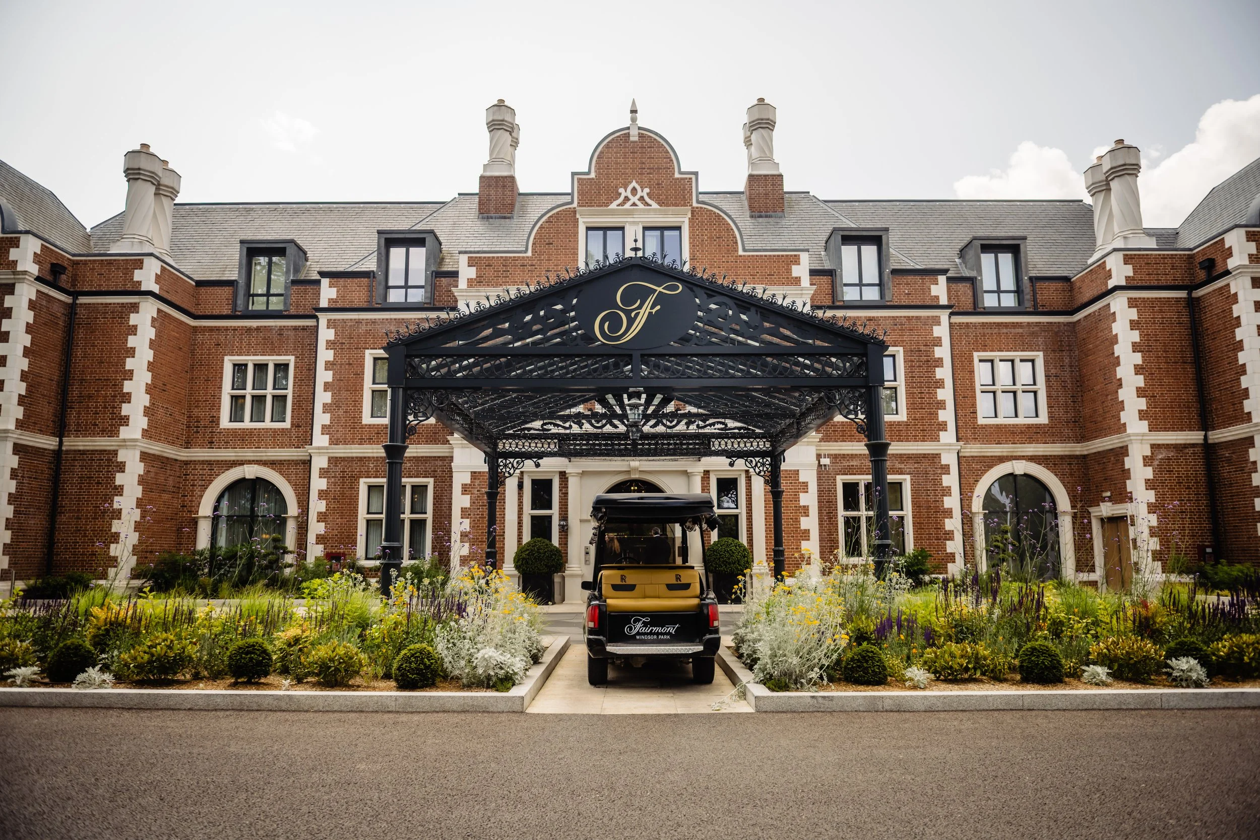 A large brick mansion with a black wrought-iron gate arching over the driveway, which has a golf cart parked beneath it. The mansion features multiple chimneys, arched windows, and intricate architectural details like white trim and decorative brickw
