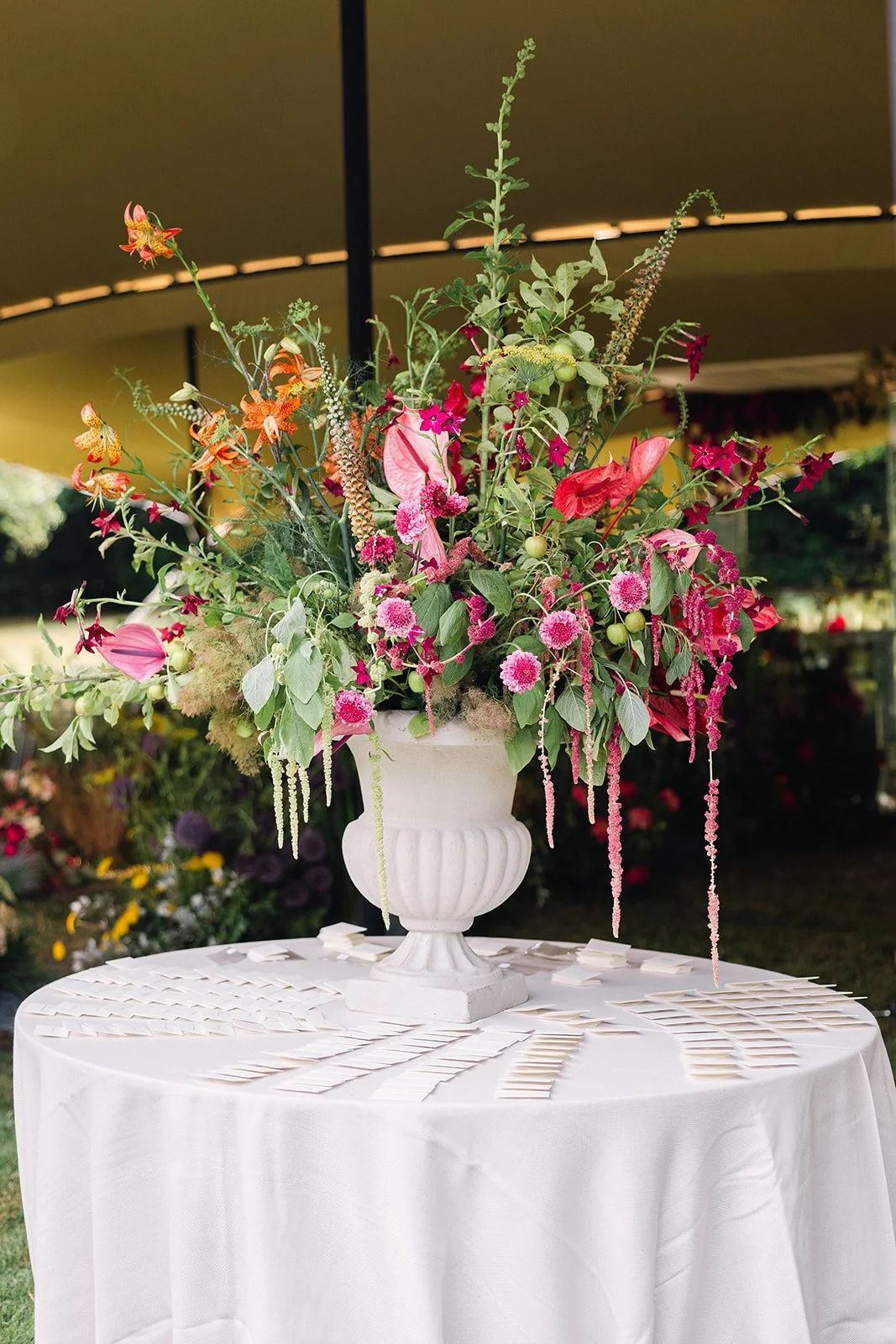 A large floral arrangement in a white urn-style vase on a white tablecloth-covered round table, outdoors, featuring pink, red, and orange flowers and greenery.