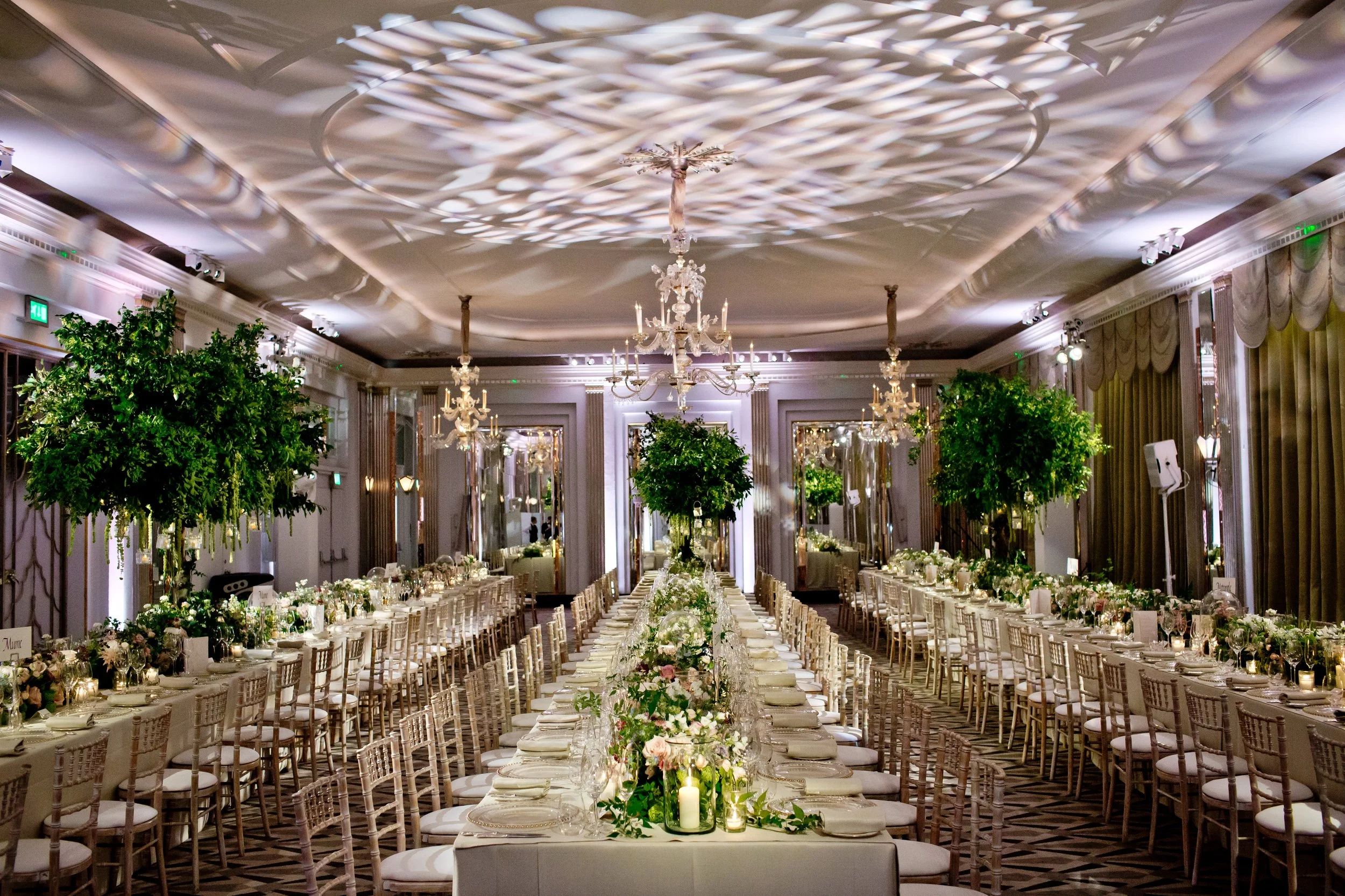 Elegant banquet hall decorated for a formal event with long tables set with silverware, glasses, and floral centerpieces, ornate chandeliers hanging from the ceiling, and greenery decor.