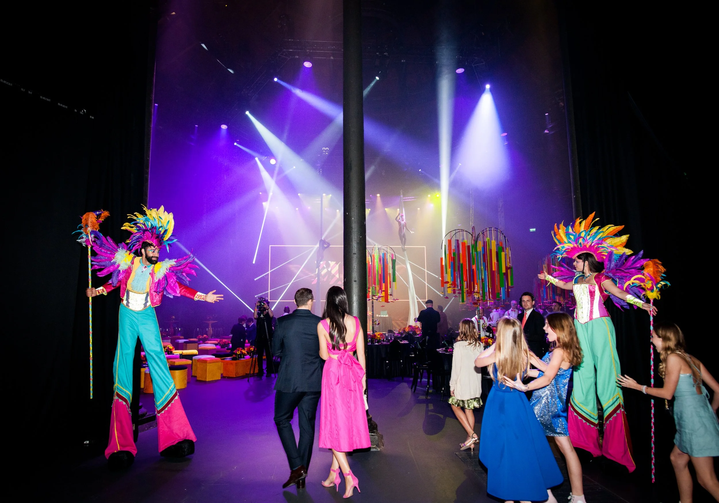 A lively stage performance with acrobats and performers in colorful costumes with feathers, set against vibrant lighting and decor, at an  Bat Mizvah the roundhouse