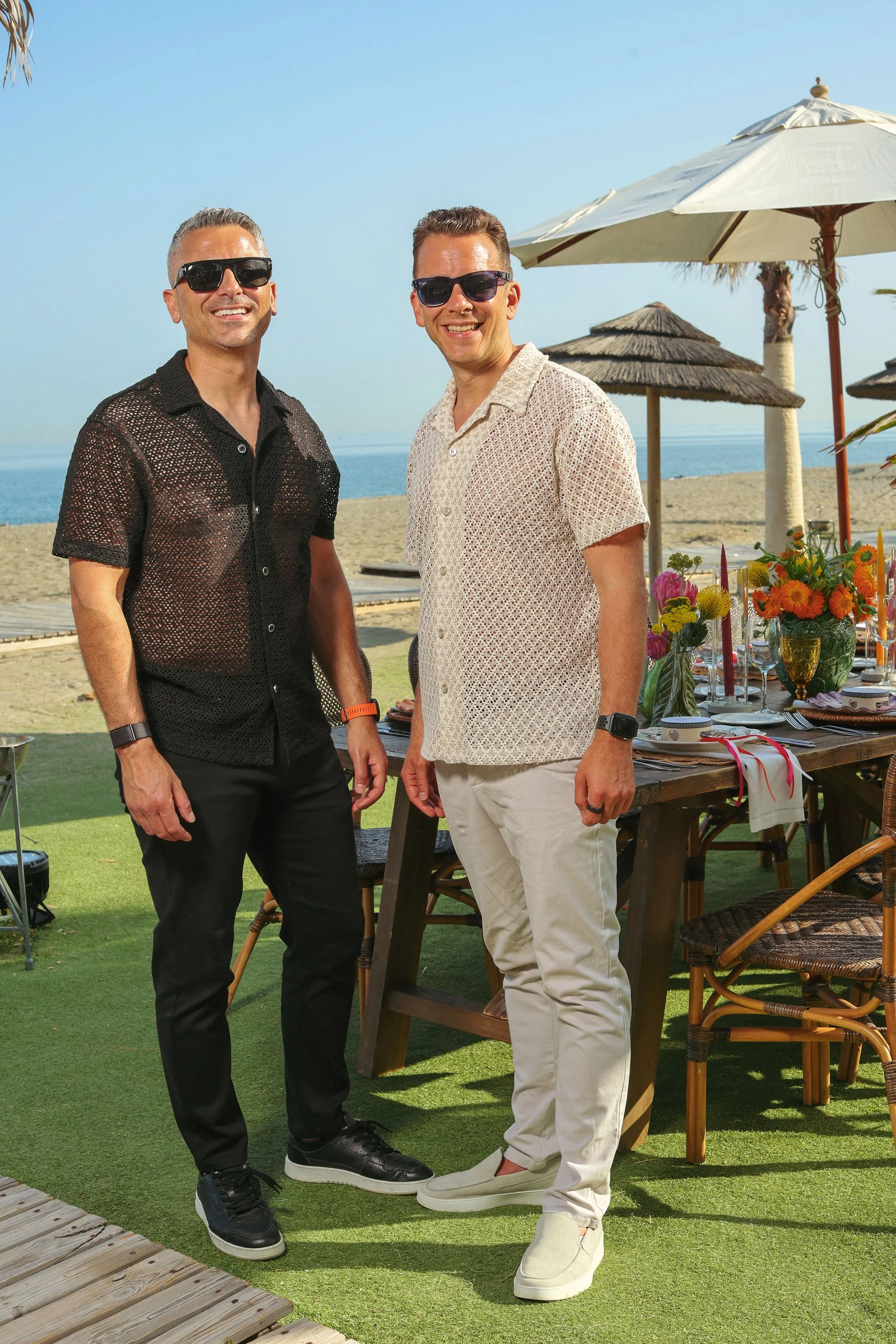 Laurie Weitzkorn and Stas Anastasiou on grassy area near the beach, smiling and wearing sunglasses. There are beach umbrellas, a table with flowers, and the ocean in the background at Anantara Villa Padierna Resort in Marbella