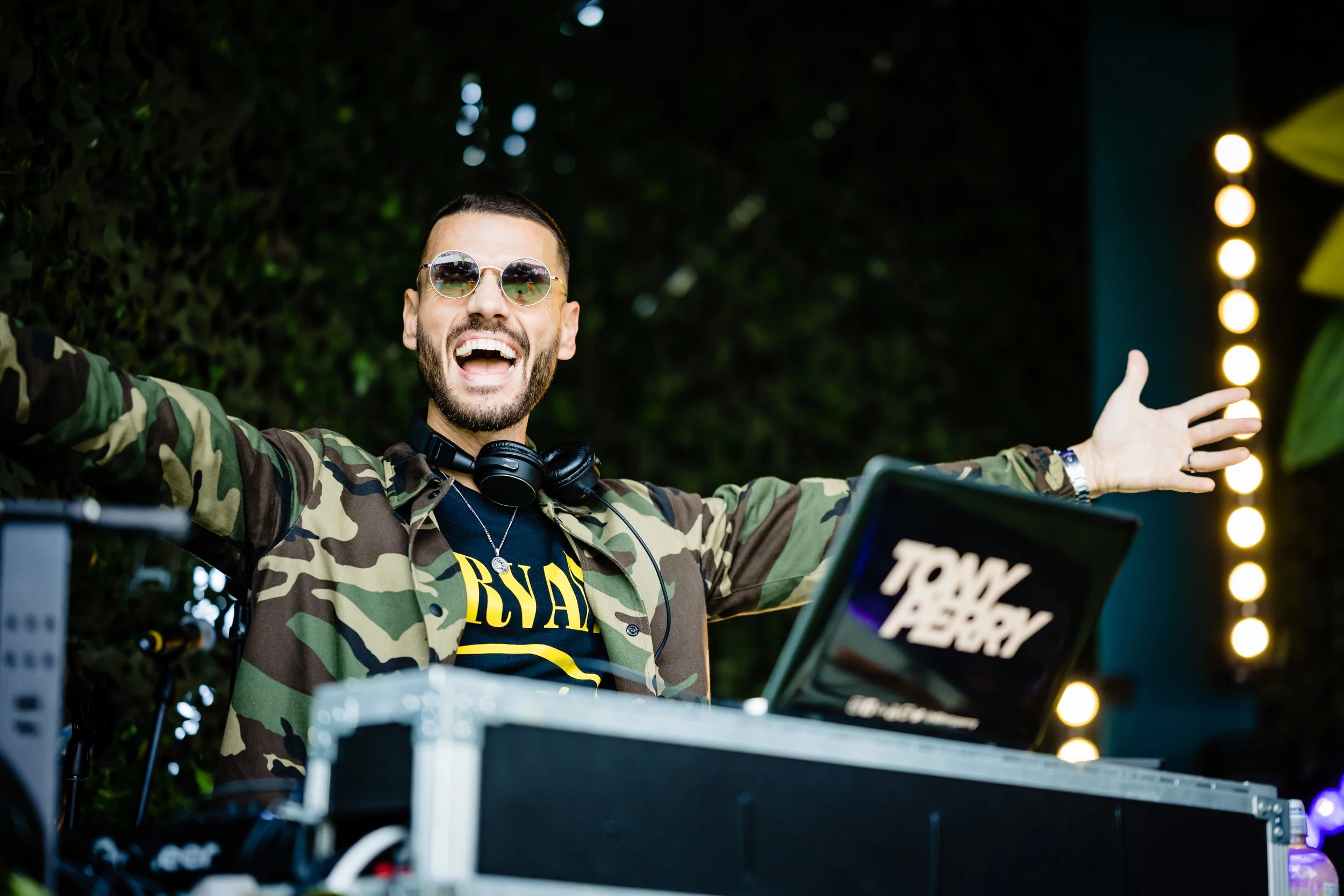 A man wearing sunglasses, a camouflage jacket, and headphones is smiling with arms outstretched in a lively outdoor setting at night, with stage lights and equipment visible at a Bar Mitzvah at  Wrotham Park. Tony Perry