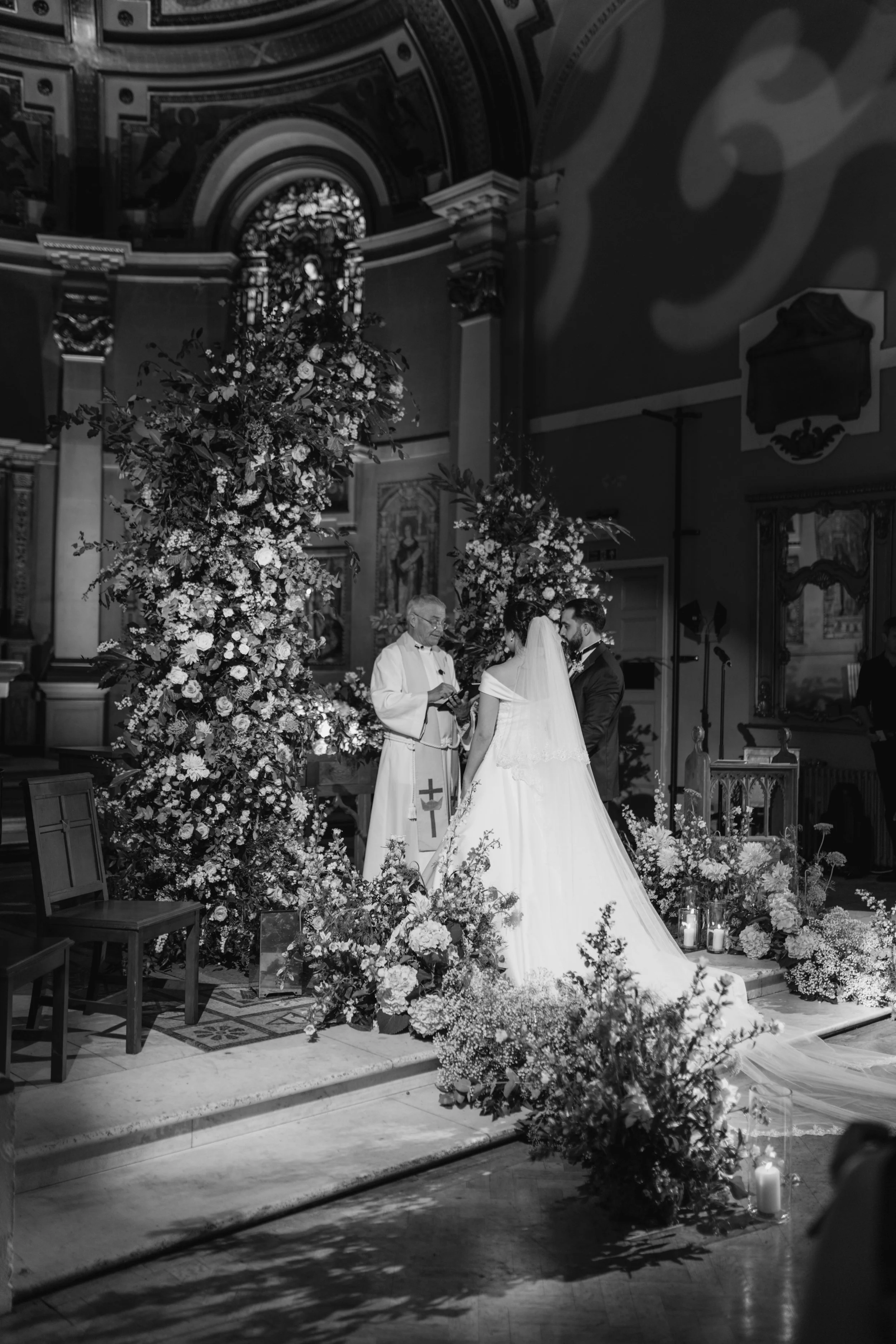 Black and white photo of a wedding ceremony inside a church, showing a bride and groom, a priest, an altar decorated with flowers and candles, and religious artwork on the walls.