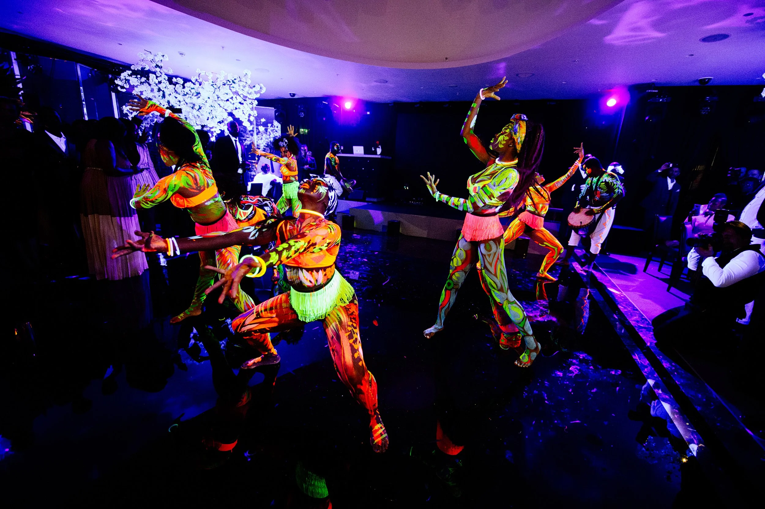People dressed in colorful, patterned outfits dancing on a dark stage with purple lighting and a decorative white tree in the background.