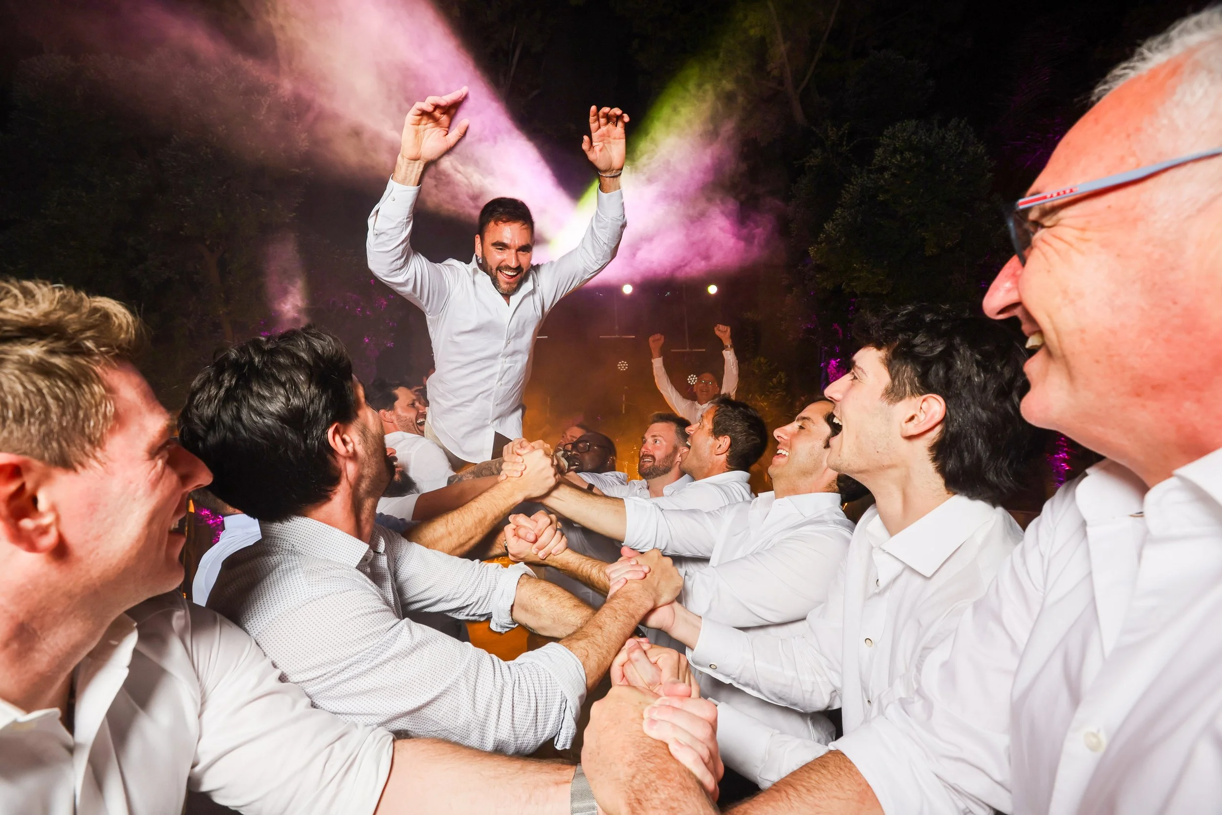 A group of smiling men in white shirts celebrating at a party surrounded by lights and colorful smoke, with a man lifted above their heads.