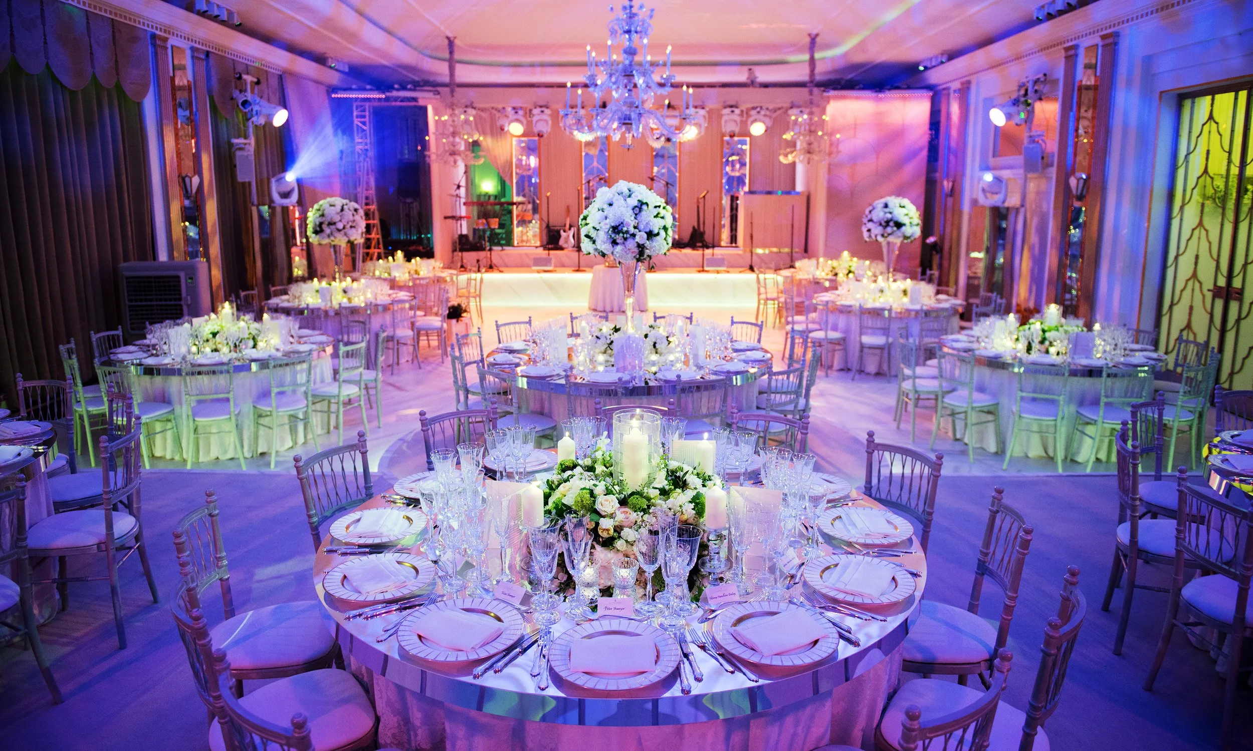 A decorated event hall with round tables set for a celebration, floral centerpieces, candles, and a stage with musical instruments at the back, illuminated with colorful lighting.