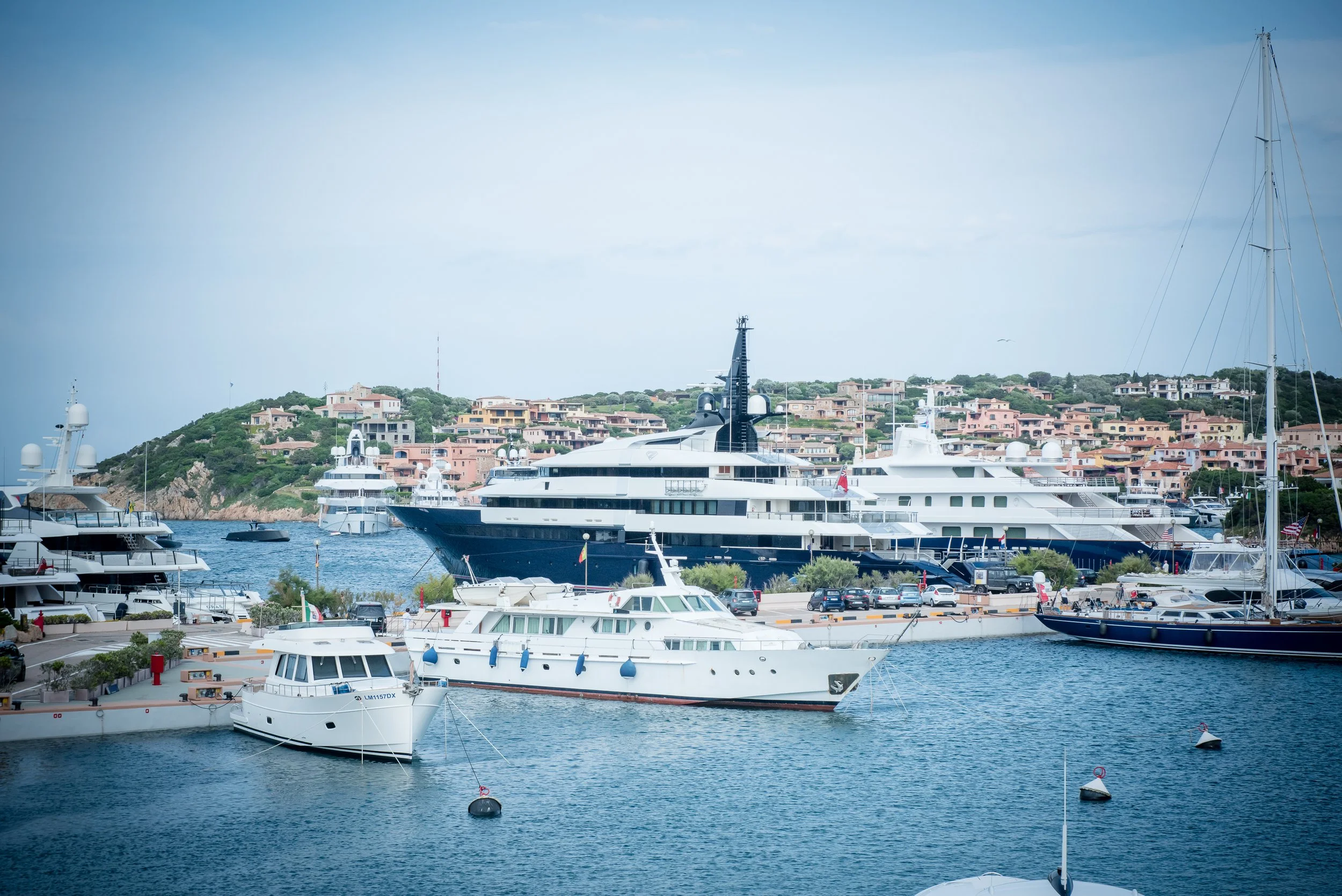 Marina with large luxury yachts docked in the water, hillside with pastel-colored houses in the background, and a clear sky.