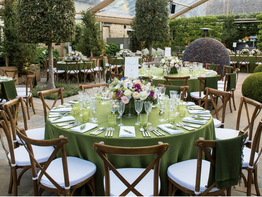 Decorated round tables with green tablecloths, floral centerpieces, glassware, and cutlery set for an event in a greenhouse or garden setting.