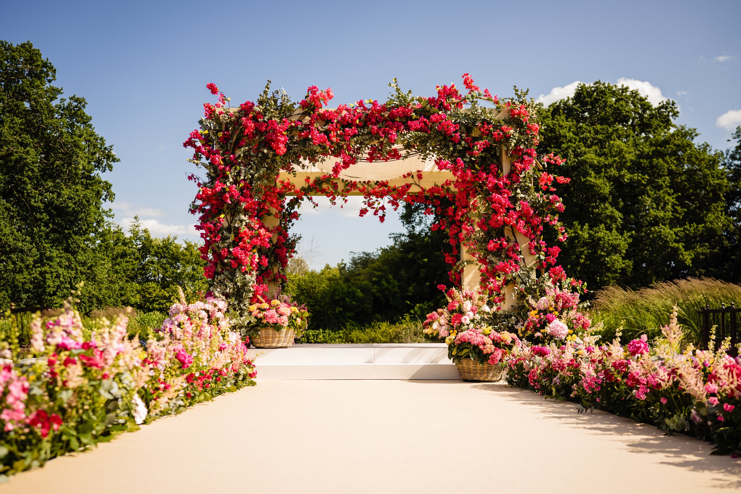 A decorated wedding ceremony arch adorned with pink and red flowers, surrounded by flower arrangements on the ground and a clear blue sky with trees in the background.Fairmont Windsor Park'.