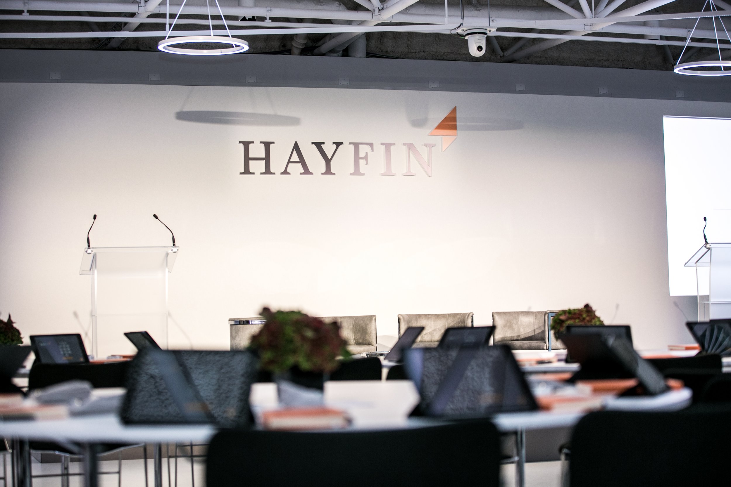 An indoor conference room with a white wall featuring the logo 'HAYFIN' and a geometric arrow design. There are microphones on stands and a row of chairs or tables in the foreground, with some potted plants and electronic devices on the tables. The c